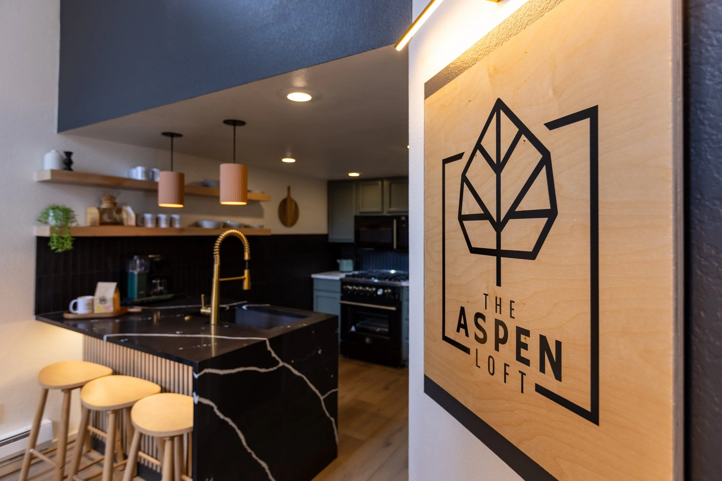 Interior view of a modern kitchen with a black marble island, gold faucet, and open wooden shelves with decorative items. A wooden sign on the wall reads 'The Aspen Loft' with a leaf logo.