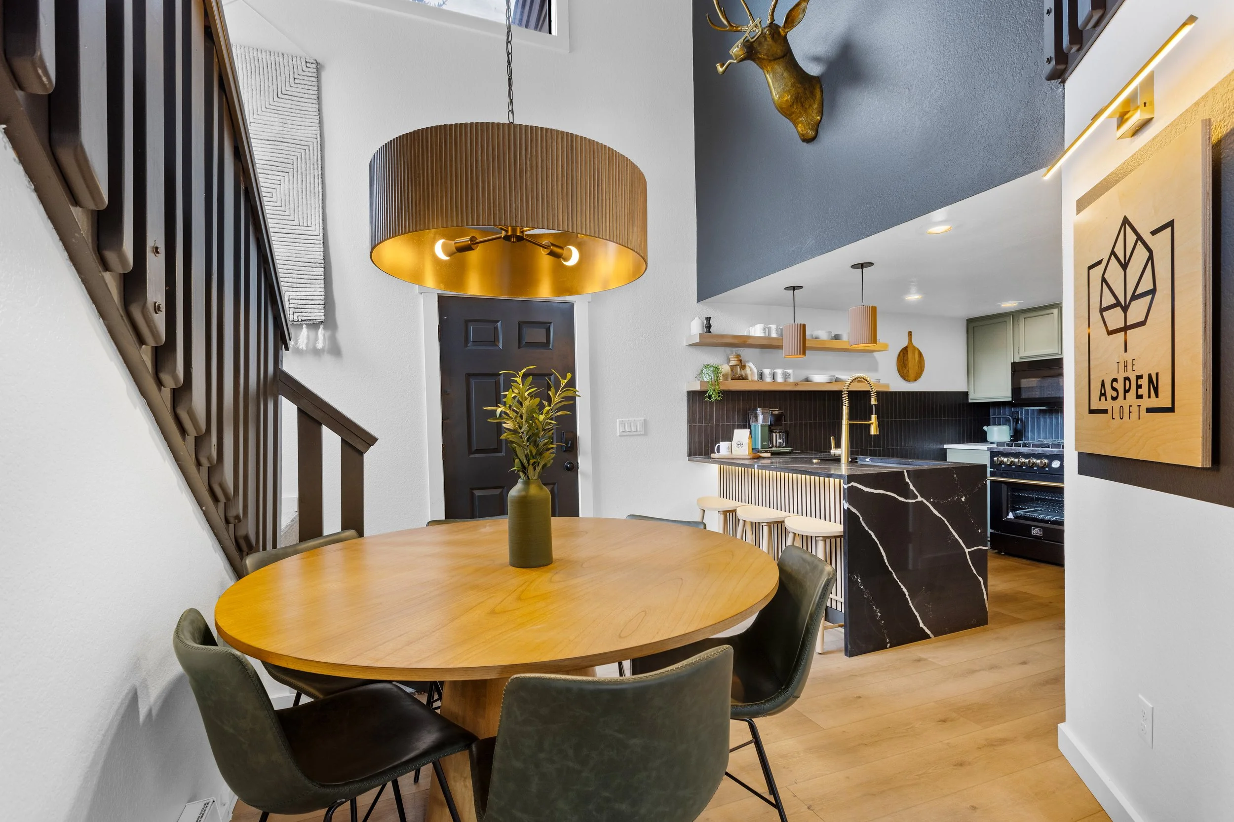 Modern kitchen and dining area with a wooden round table, black and green chairs, a black front door, pendant lights, and a black marble kitchen island.
