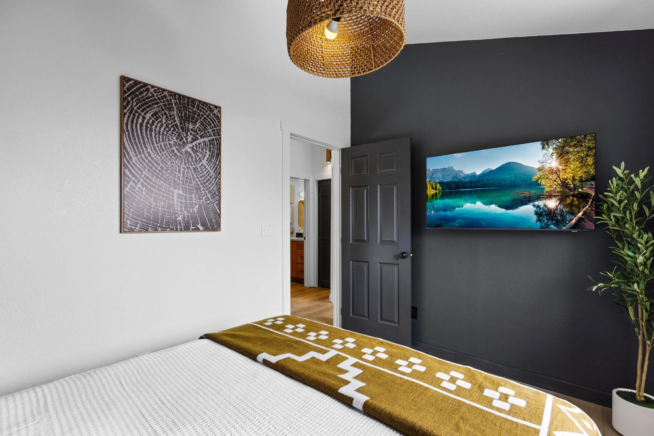 A bedroom with white and dark gray walls, a wooden light fixture hanging from the ceiling, a framed art print of a tree ring pattern on the wall, a flat-screen TV displaying a nature scene, and a potted plant in the corner. Part of a bed with a white
