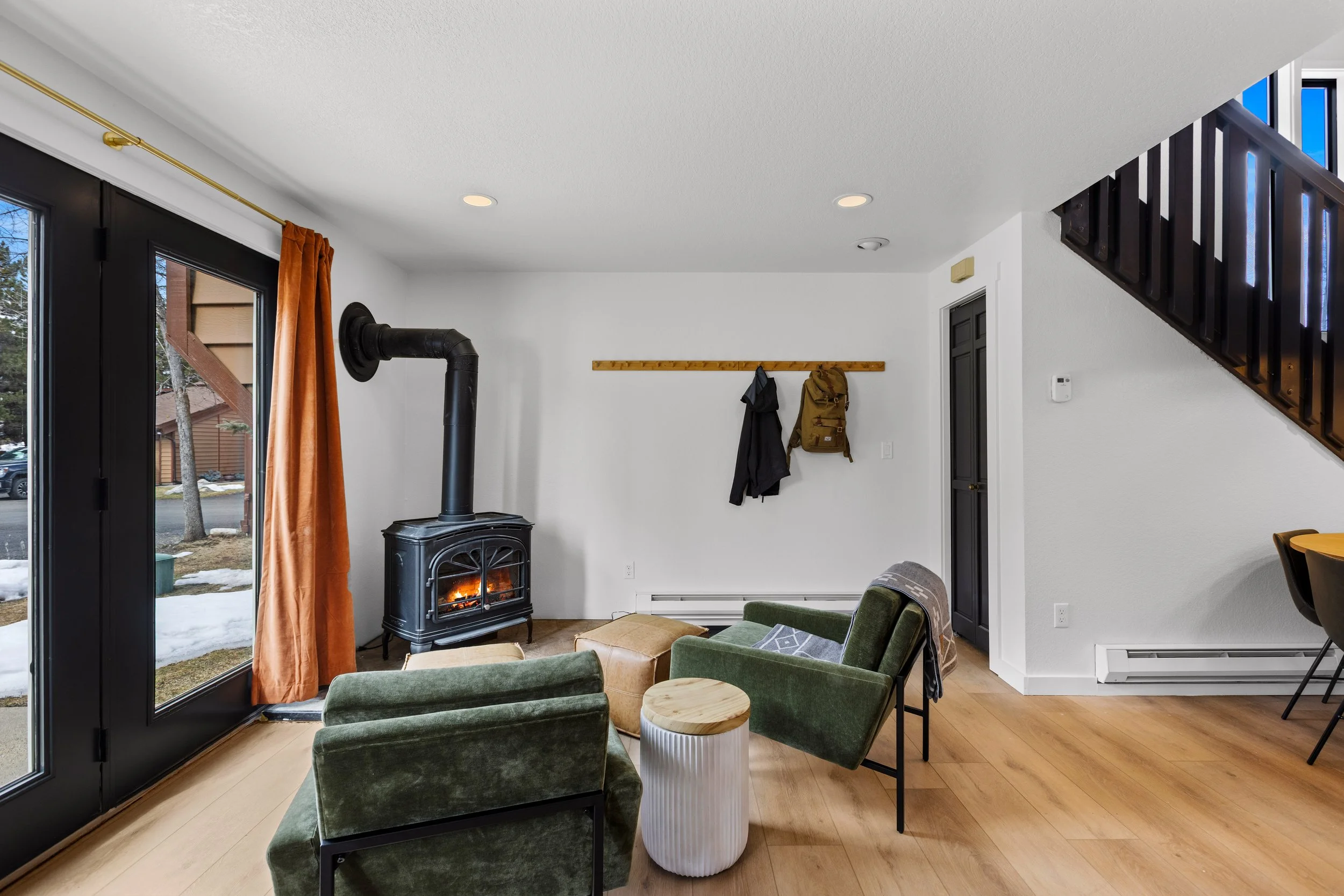 Living room with a wood stove, green armchairs, a small round white side table, sliding glass door with orange curtains, wall hooks with jackets and backpack, and staircase railing.