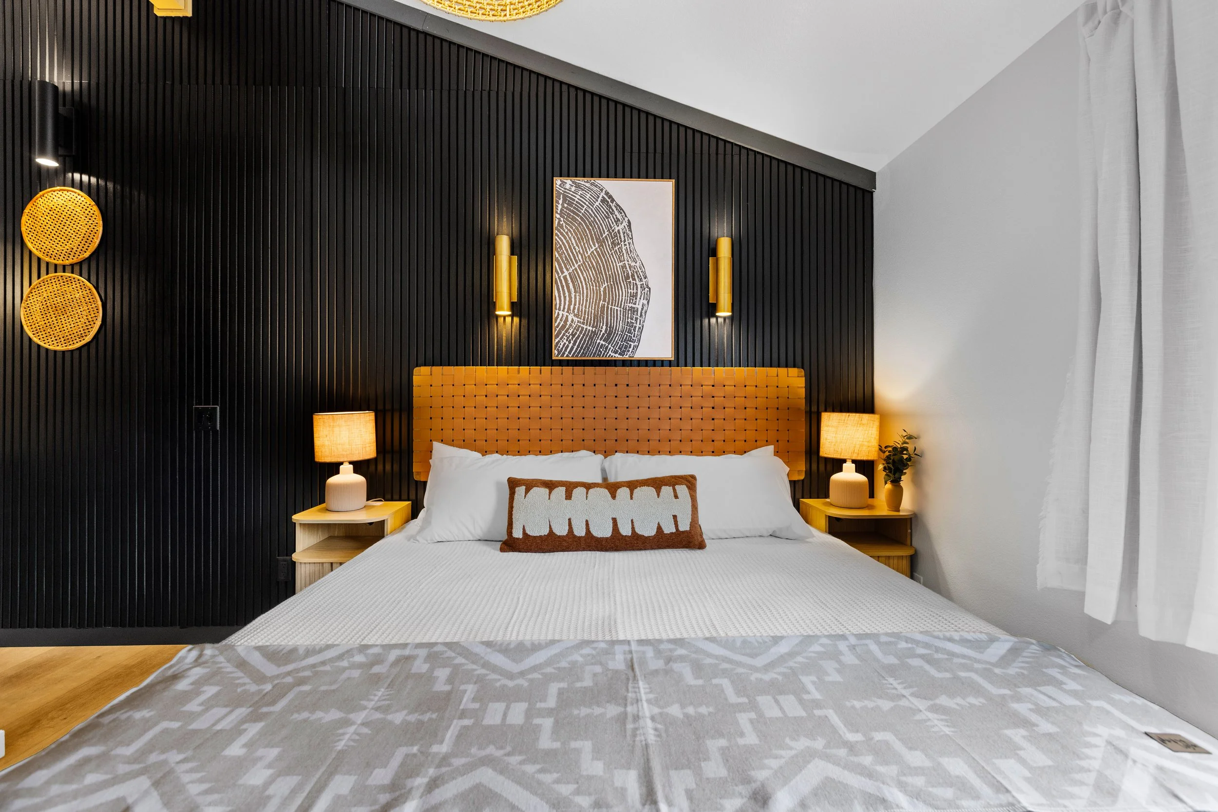 A neatly made bed with white bedding and a decorative brown pillow in a modern bedroom with black and white walls. The bed has an orange headboard, flanked by matching yellow nightstands with lamps. There is artwork and wall-mounted lights above the 