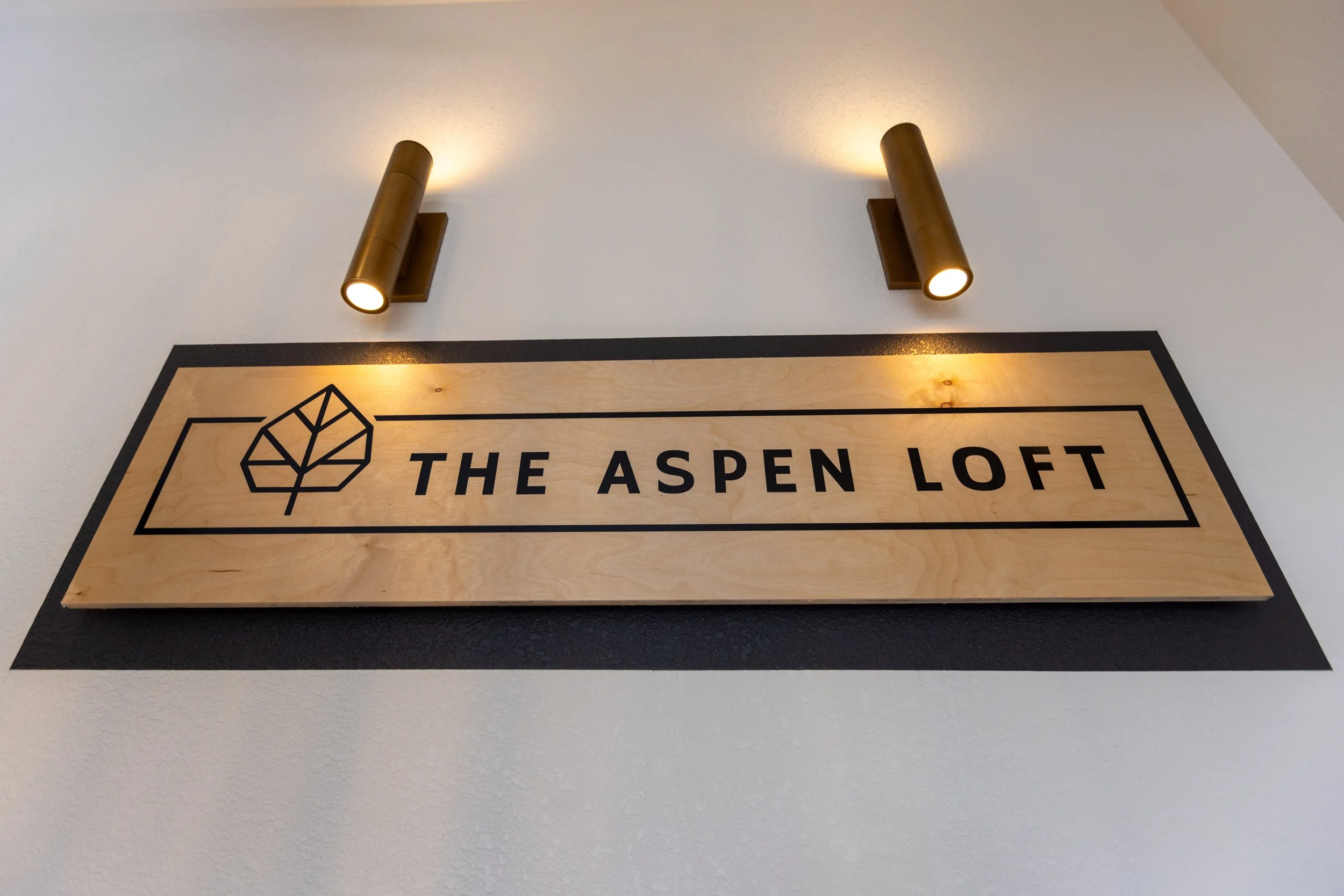 Wooden sign that reads "The Aspen Loft" with a simple leaf logo above the text, illuminated by two brass wall-mounted lights.