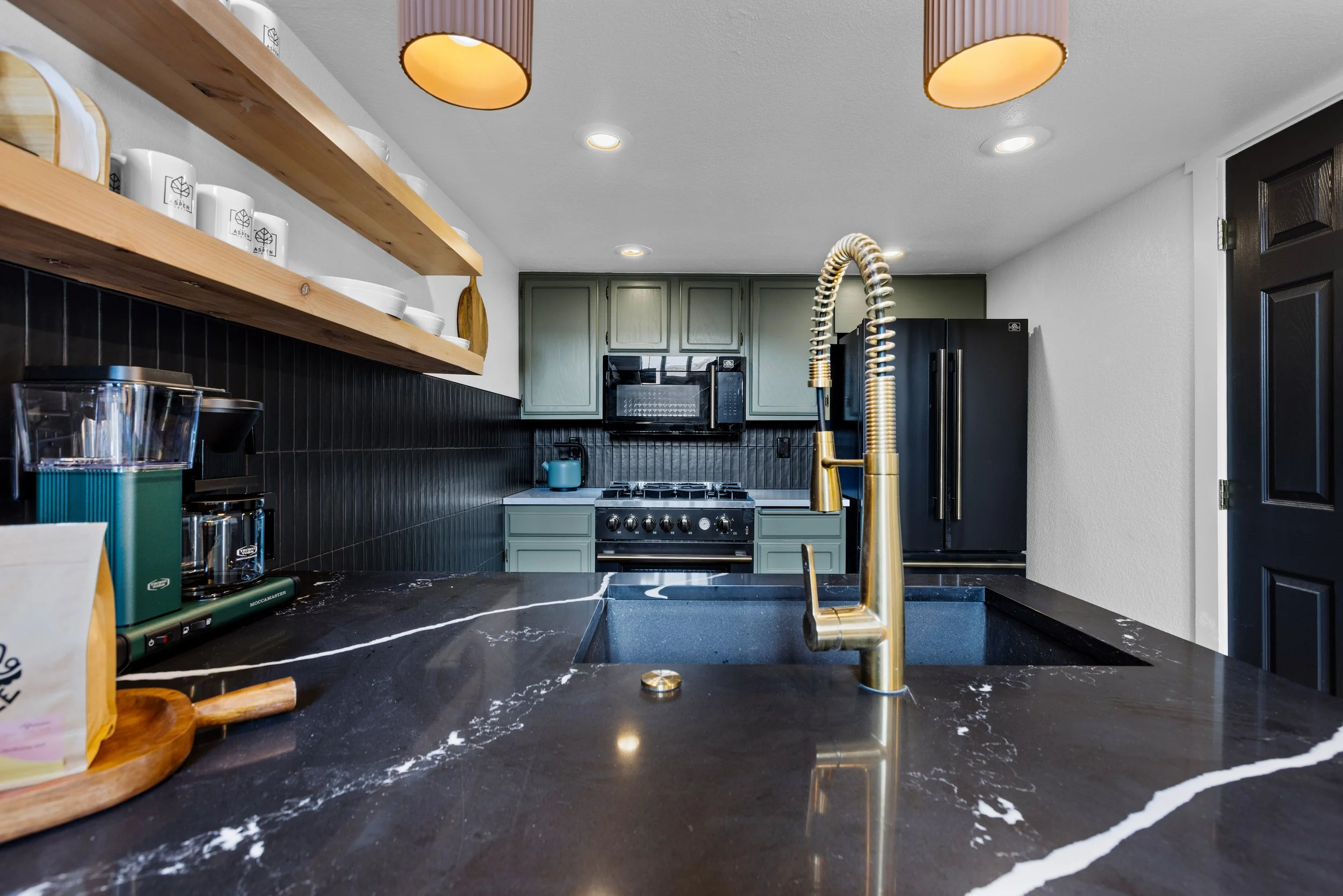 Modern kitchen with black marble countertop, gold faucet, green cabinets, black refrigerator, and open wooden shelves with dishes.