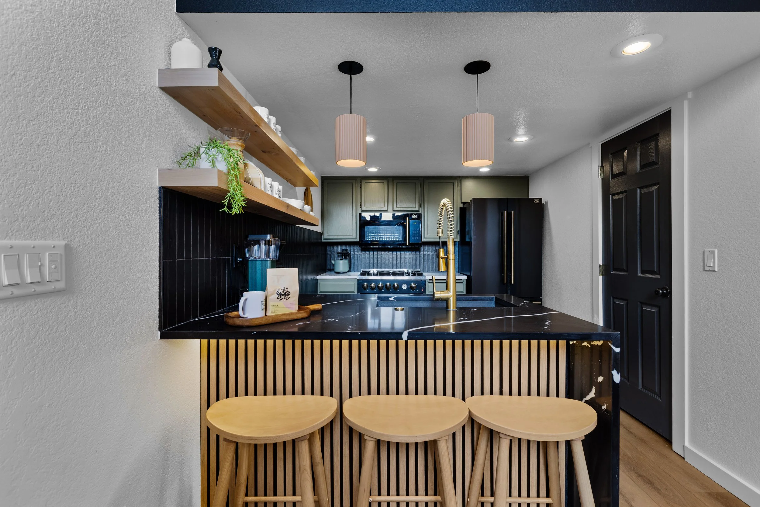 Kitchen with black countertops and backsplash, green cabinets, gold fixtures, black refrigerator, open wooden shelves with decor, and three light fixtures hanging above the island with three wooden stools.