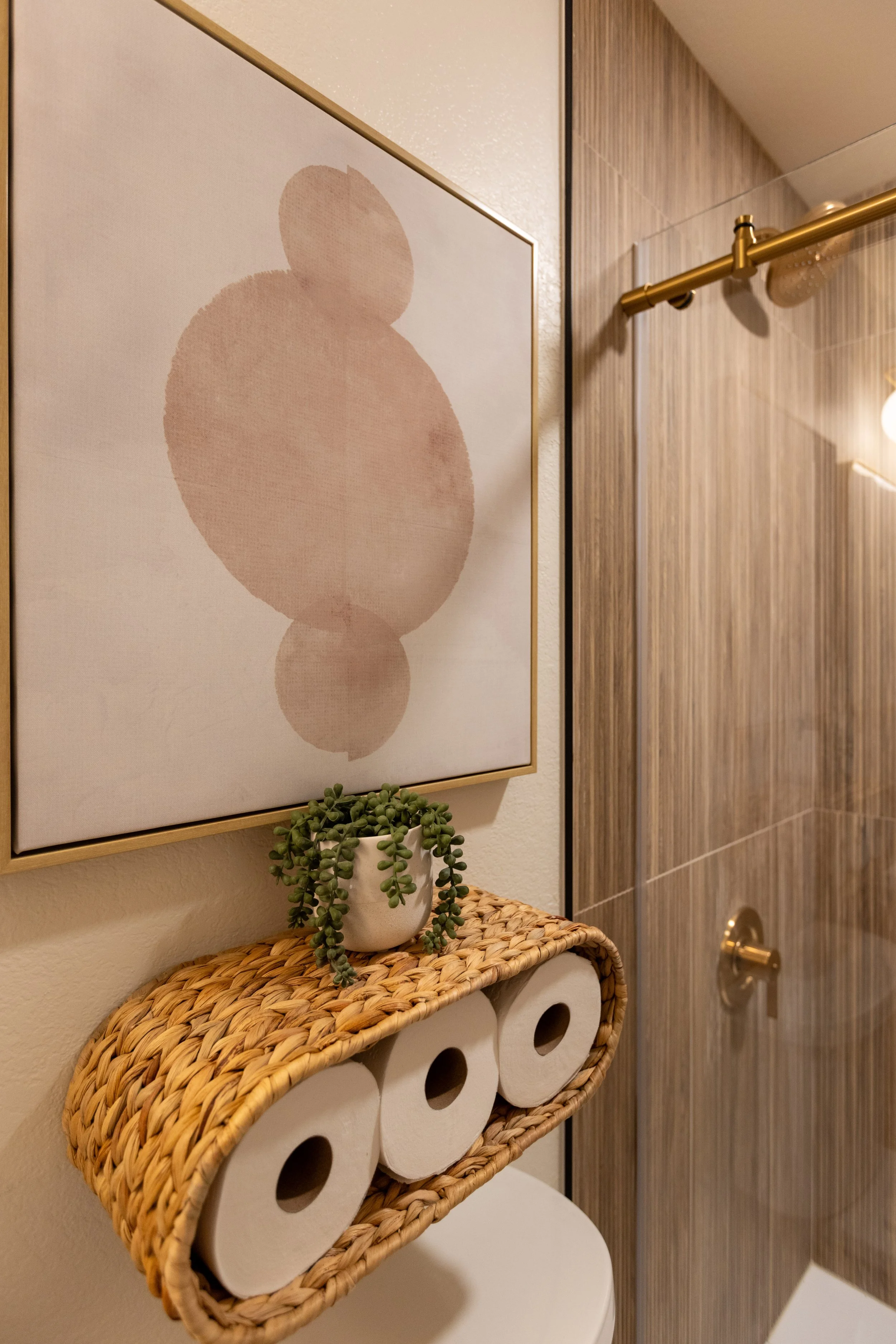 Bathroom with wicker storage shelf holding three rolls of toilet paper and a small potted plant, with a framed abstract art piece on the wall and a shower area with wood-like tiles and gold fixtures.