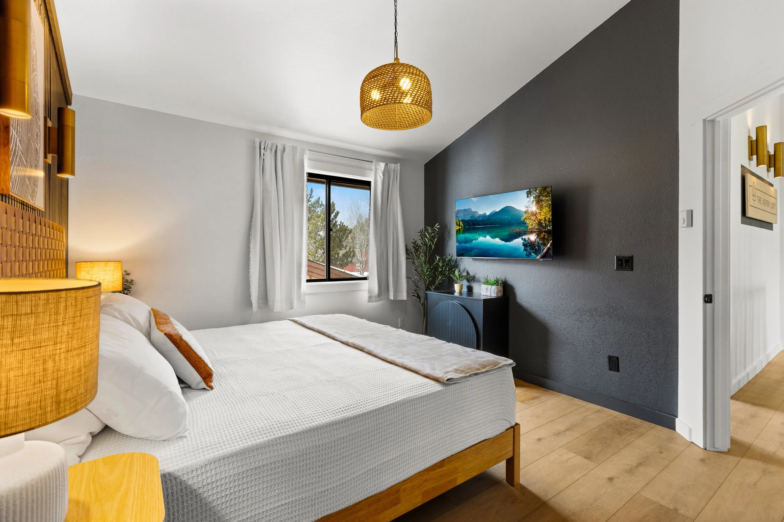 A modern bedroom with a bed, nightstand with lamp, a TV mounted on a dark accent wall, and a window with white curtains showing trees outside. The room has wooden flooring and contemporary decor.