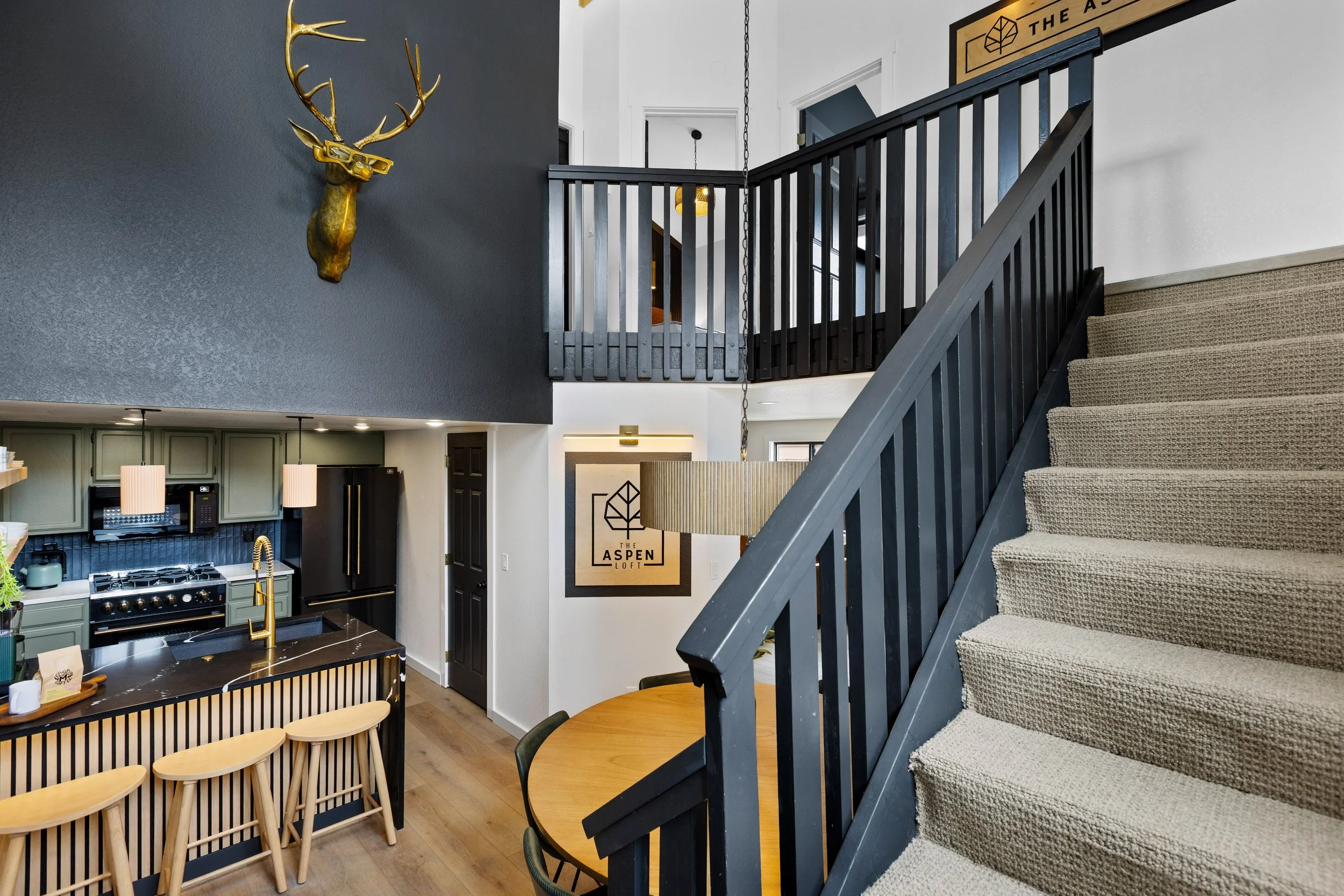 Interior view of a modern multi-story home with a staircase, kitchen, and dining area. The staircase has a dark gray railing and beige carpeted steps. The kitchen features green cabinets, a black island with a gold faucet, and stools. A decorative go