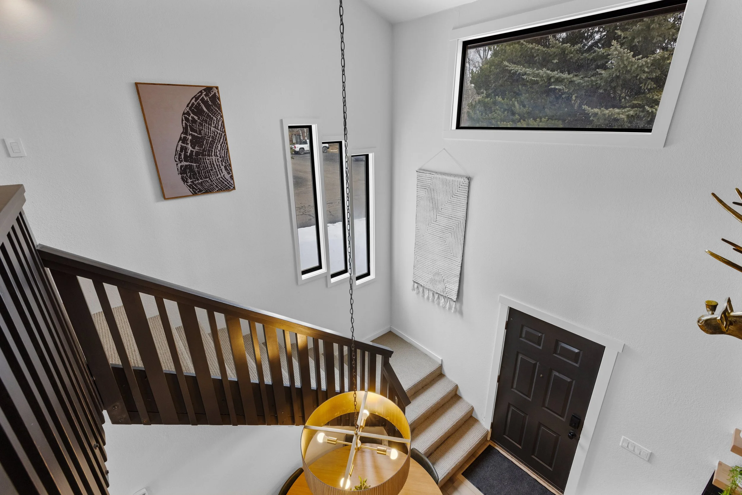 Interior view of a house entryway with stairs, black front door, large horizontal window, and white walls decorated with artwork and a wall hanging, taken from the upper level.