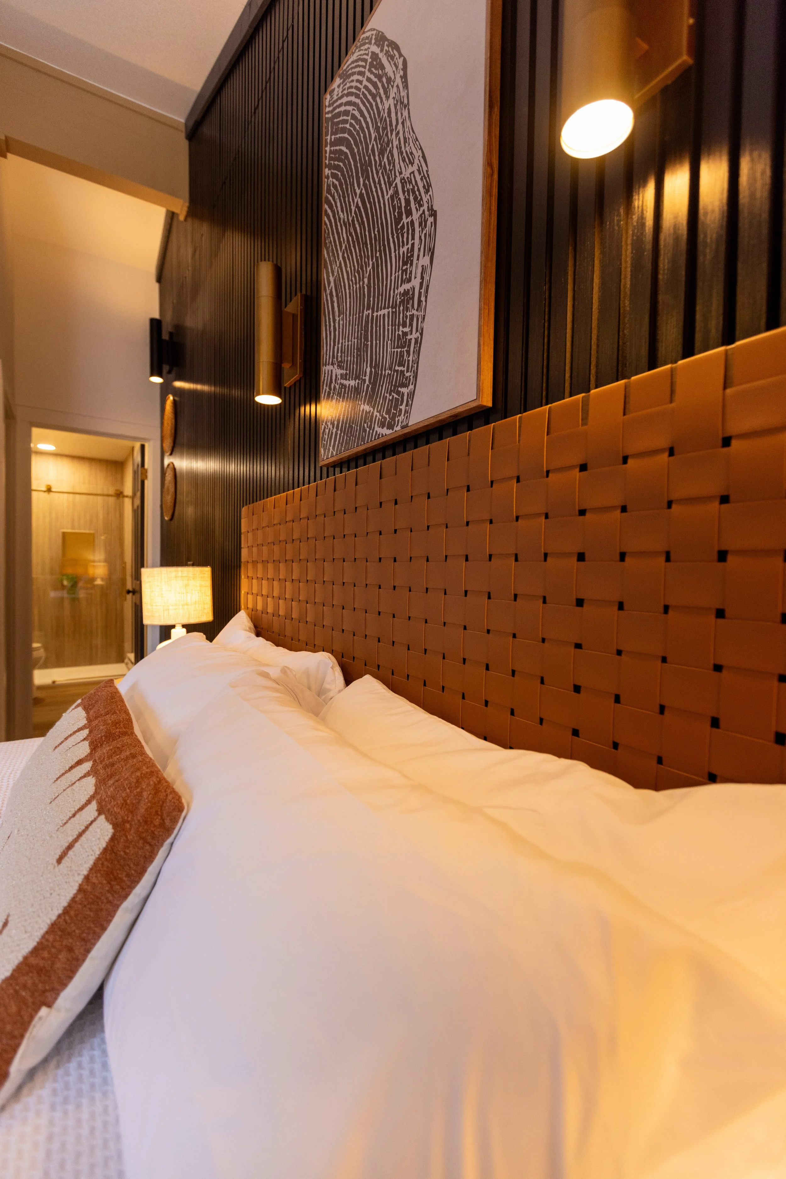 A cozy bedroom with a large bed, white pillows, and an orange woven headboard. There is a lamp on the bedside table, a piece of wall art above the bed, and warm lighting throughout the room.