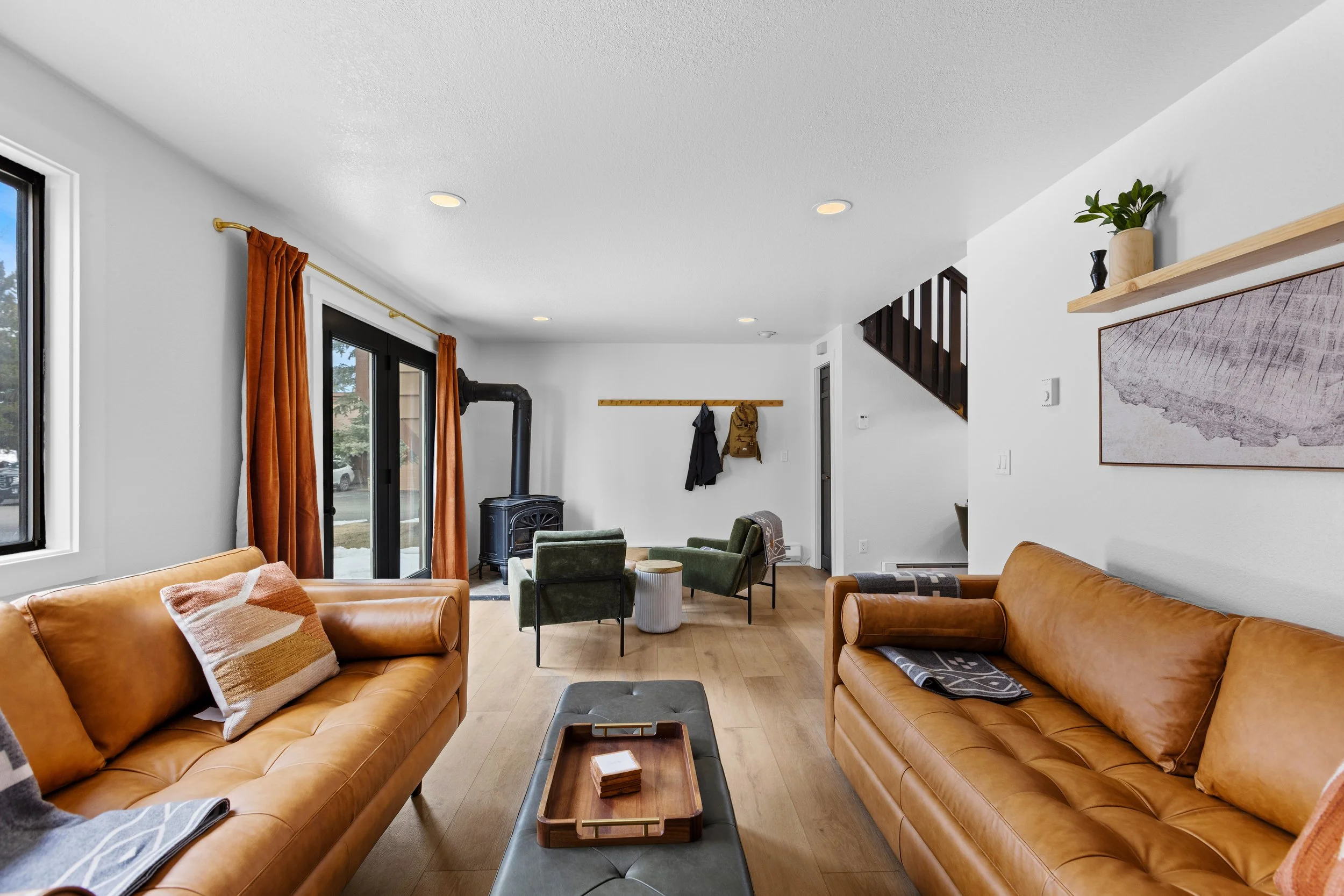 Living room with two tan leather sofas, a wooden coffee table, green armchairs near a black stove, and large windows with orange curtains.