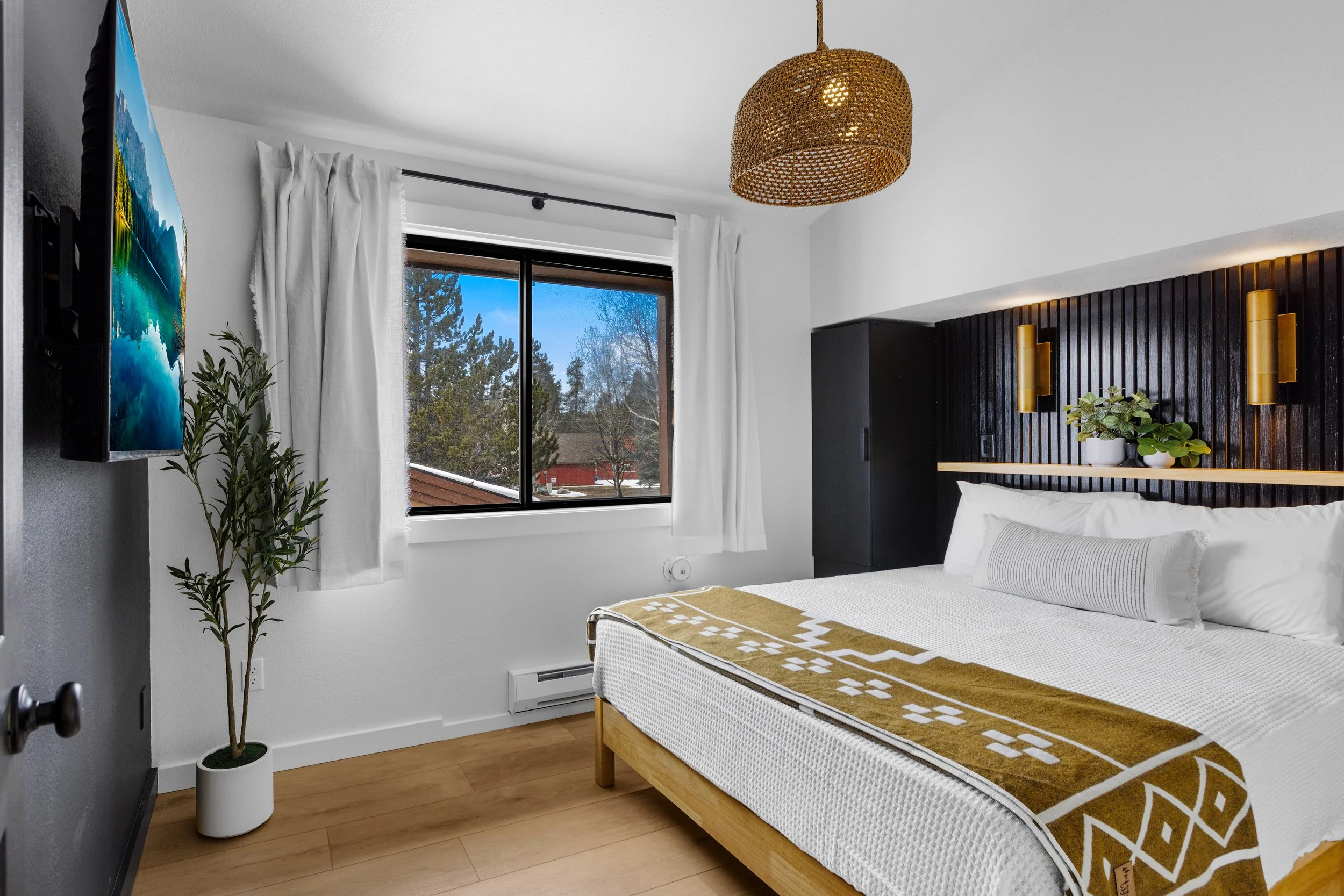 A modern bedroom with a window showing trees outside, a mounted TV, and a bed with white bedding and a patterned blanket. There is a black wood-paneled headboard with plants and wall sconces, a rattan ceiling light, and a potted plant in a white pot.
