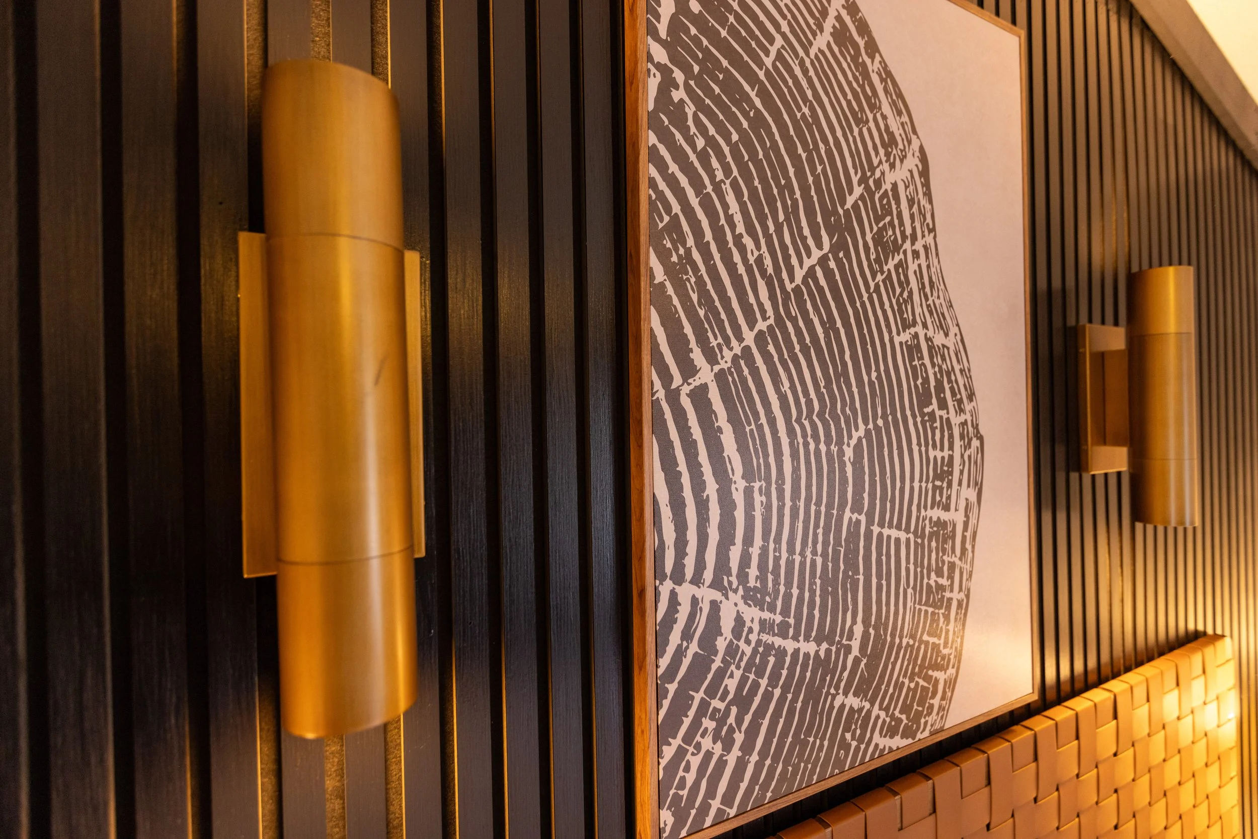 Interior wall decor featuring black vertical wood slats, two gold cylindrical wall sconces, a framed graphic art piece with an abstract pattern, and part of a mustard-colored cushioned seating area.