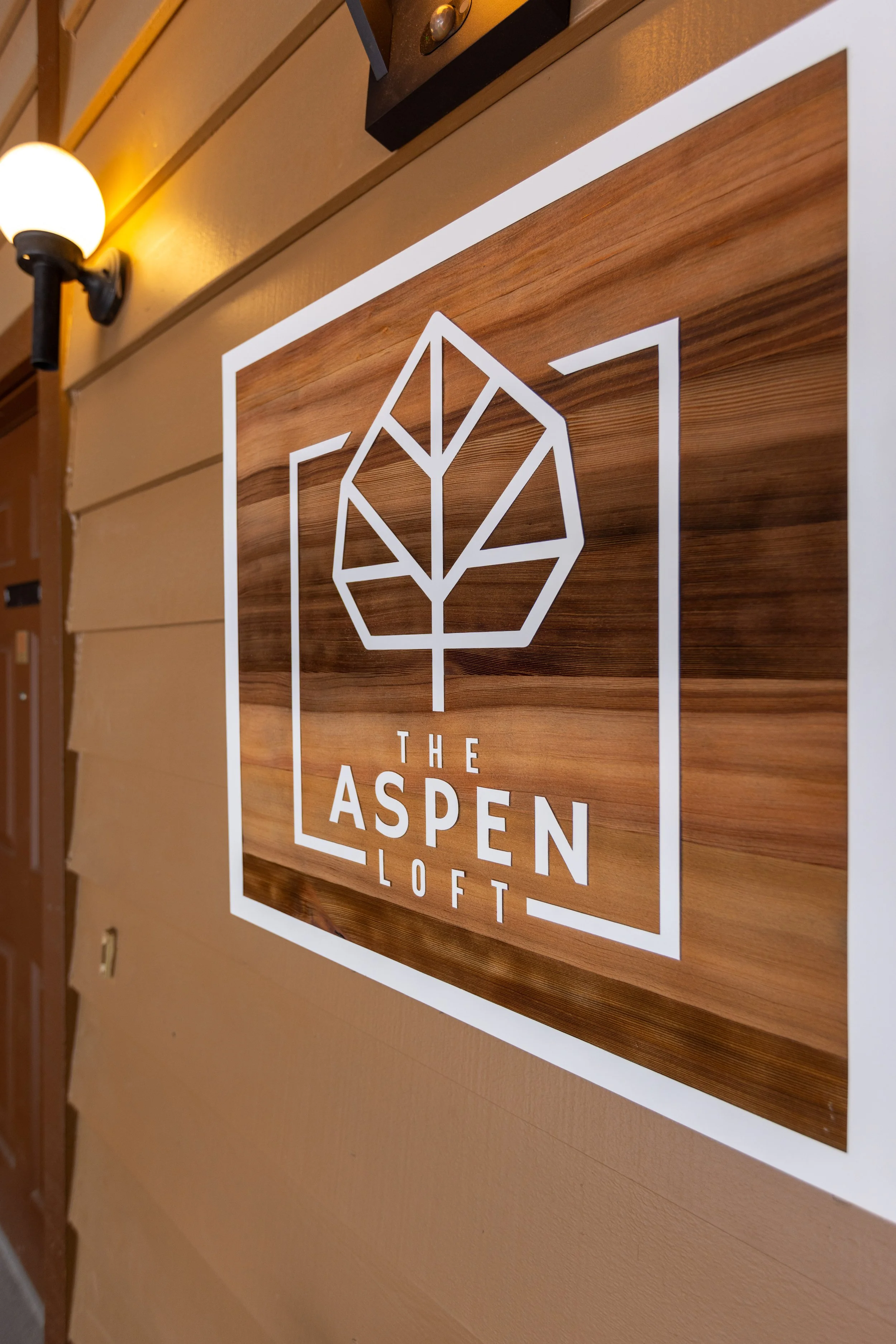 Wooden sign with white geometric leaf logo and text 'The Aspen Loft' on a wooden-paneled wall, illuminated by a wall-mounted lamp.