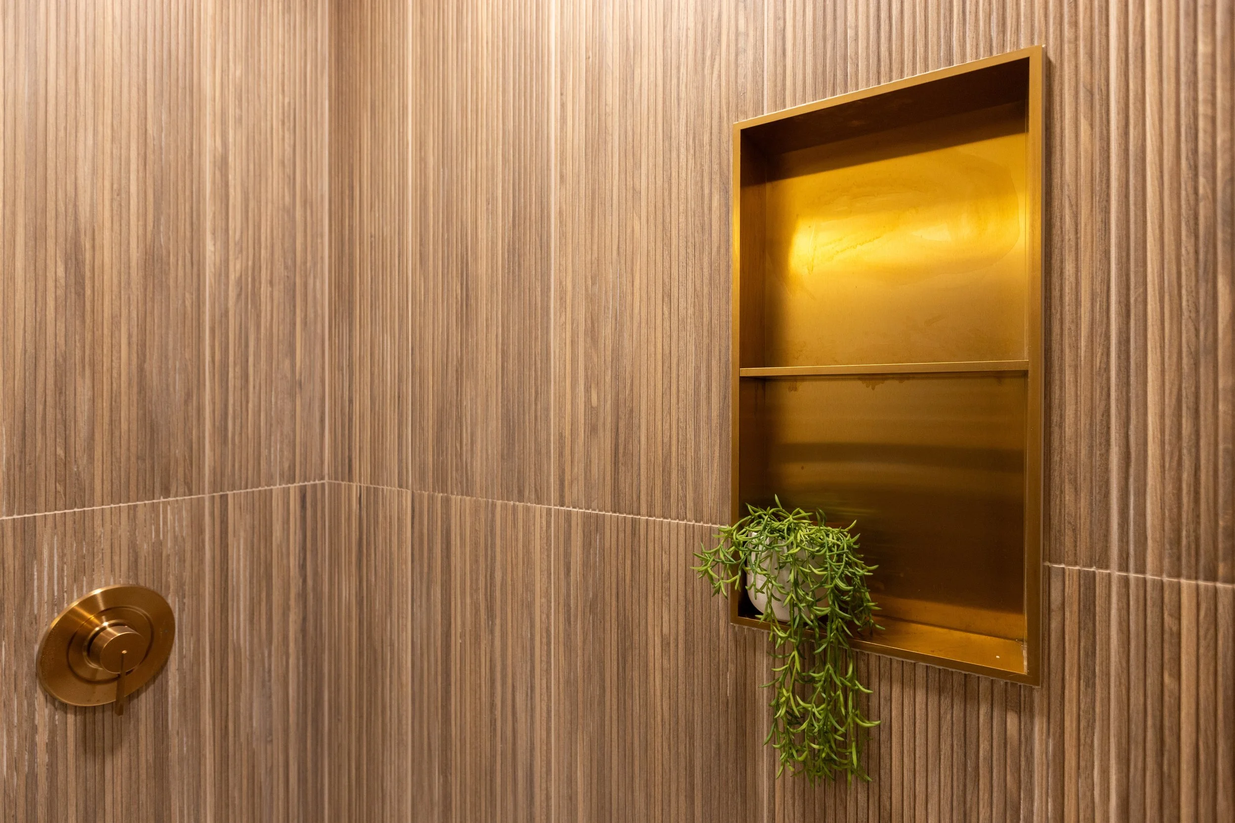 A bathroom with wood-paneled walls, featuring a built-in recessed shelf with a potted plant, and a gold-colored shower control knob.