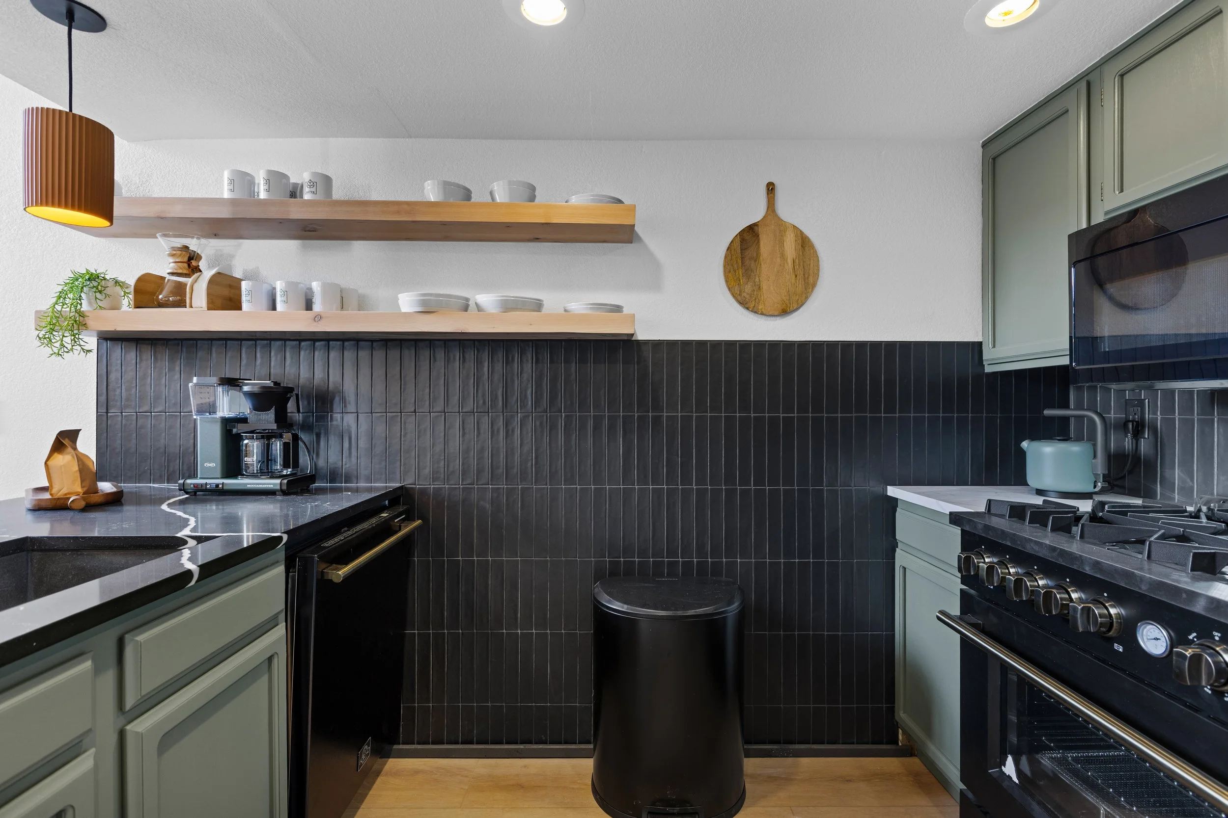 Modern kitchen with light green cabinets, black tiled backsplash, gray countertop, black stove, coffee maker, kettle, and wooden open shelves holding cups, bowls, and decorative items, with a wooden cutting board hanging on the wall.