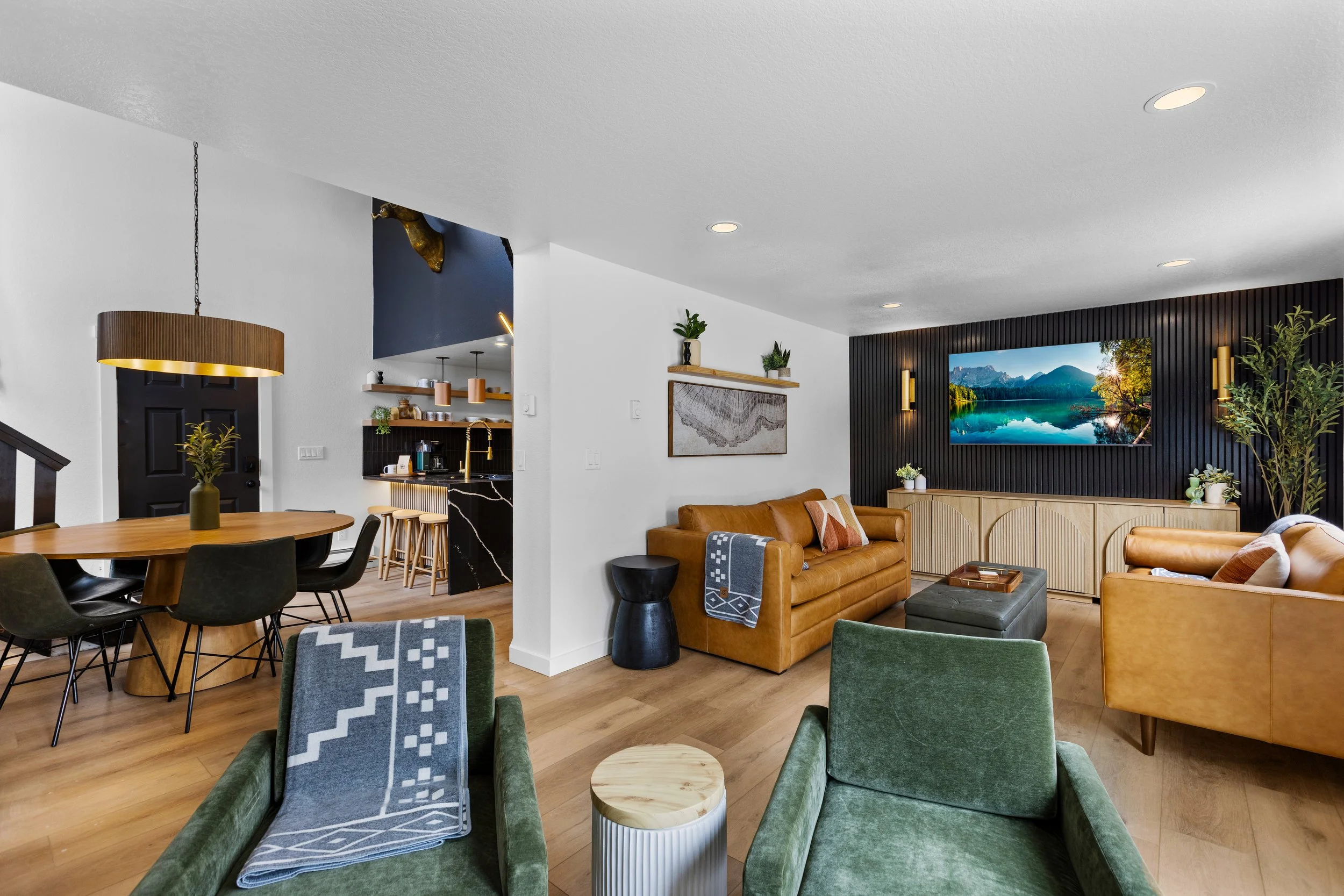 Modern living room with leather sofas, a wall-mounted TV, green velvet armchairs, a dining table with black chairs, and a kitchen area in the background