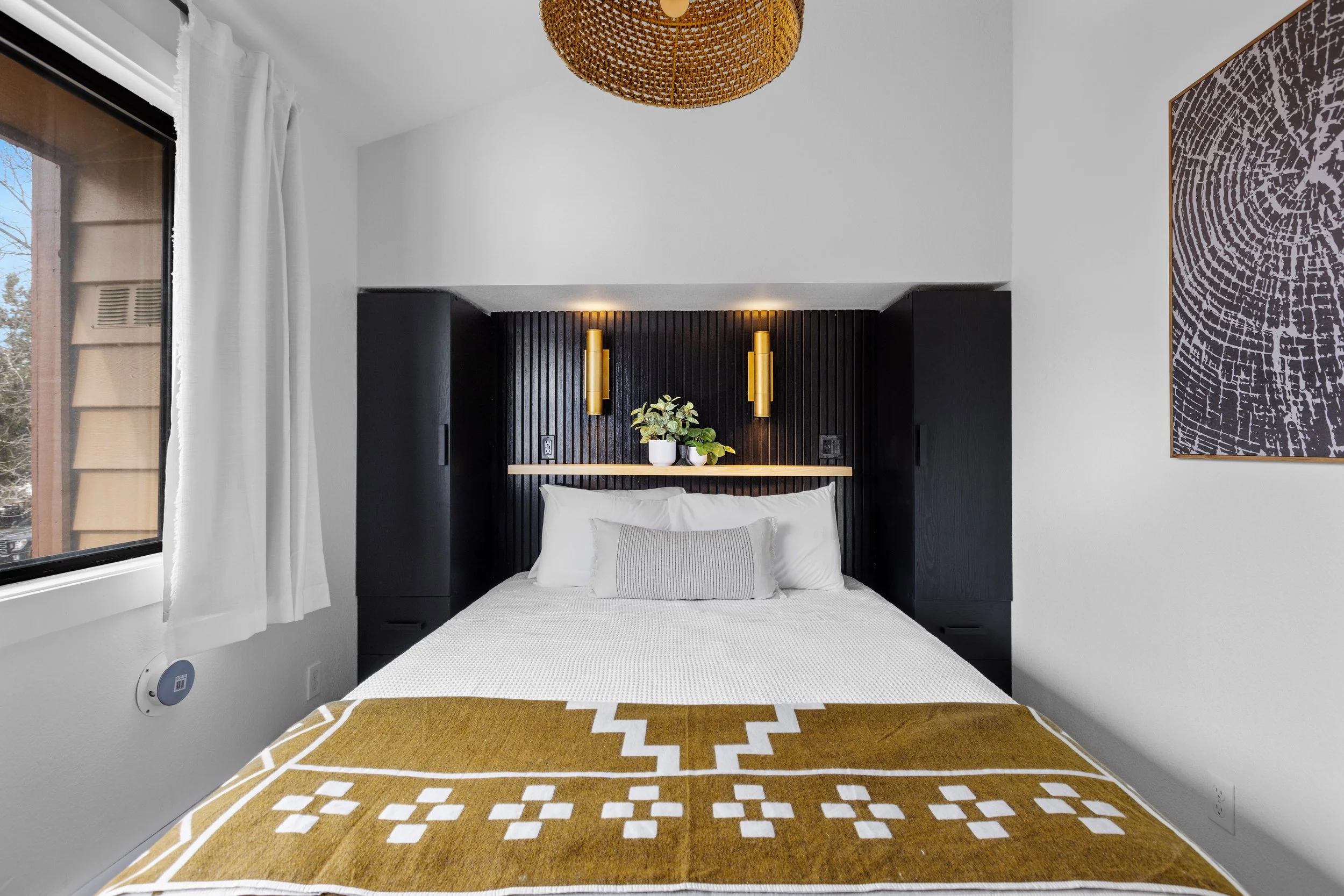 Modern bedroom with a bed featuring white bedding and a mustard yellow blanket, centered in front of a black wood-paneled headboard with wall sconces and decorative plant, white walls, art on right, window with white curtains on left.