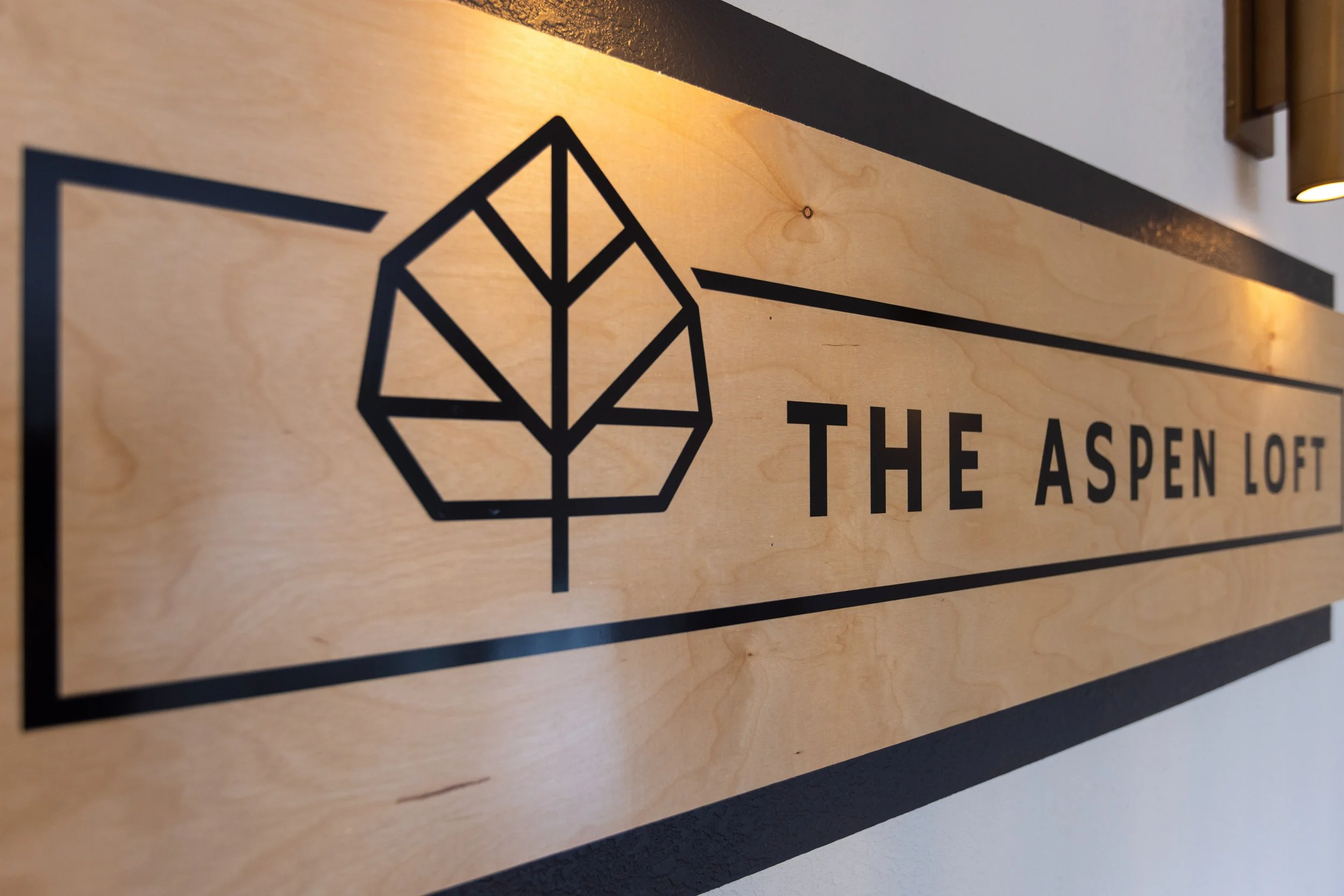 Wooden sign with black geometric leaf logo and text reading 'The Aspen Loft'.