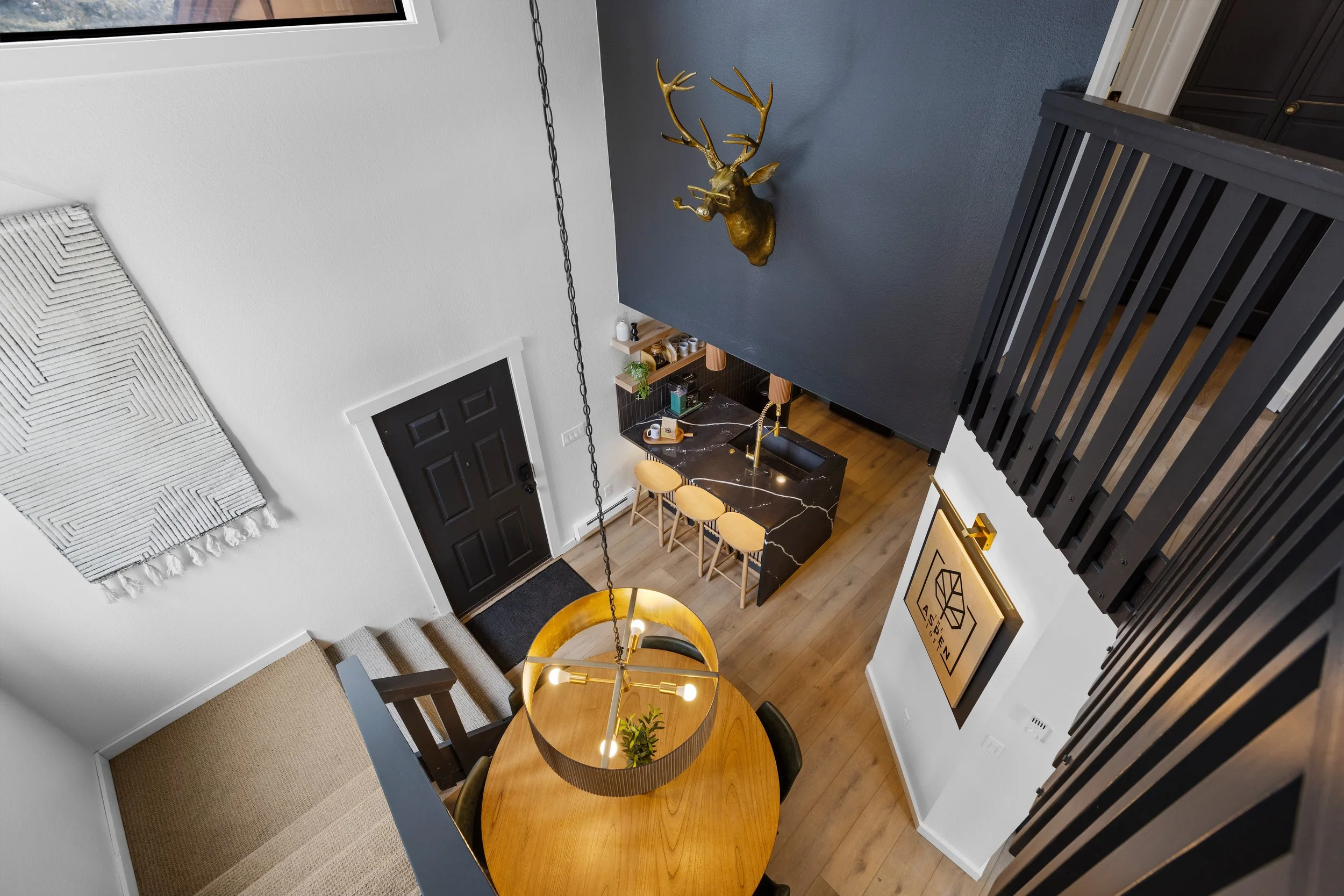 An overhead view of a modern living space with a wooden dining table, a black and gold chandelier, a breakfast bar with yellow stools, a black door, artwork on the walls, and a golden deer head mounted on a dark wall.