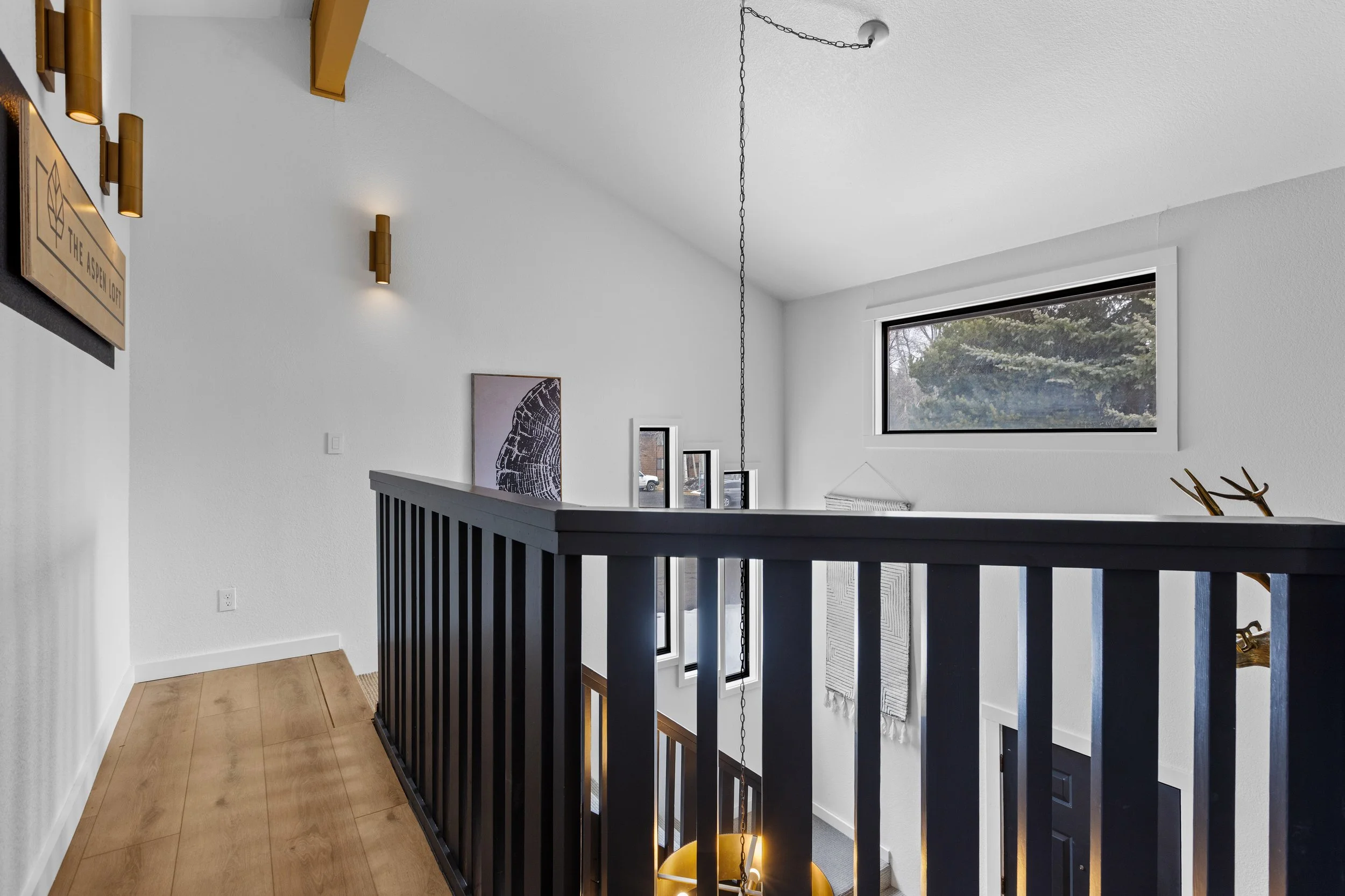 Interior view of a modern staircase landing with white walls, black railing, light wood floor, a window letting in natural light, and decorative wall art.