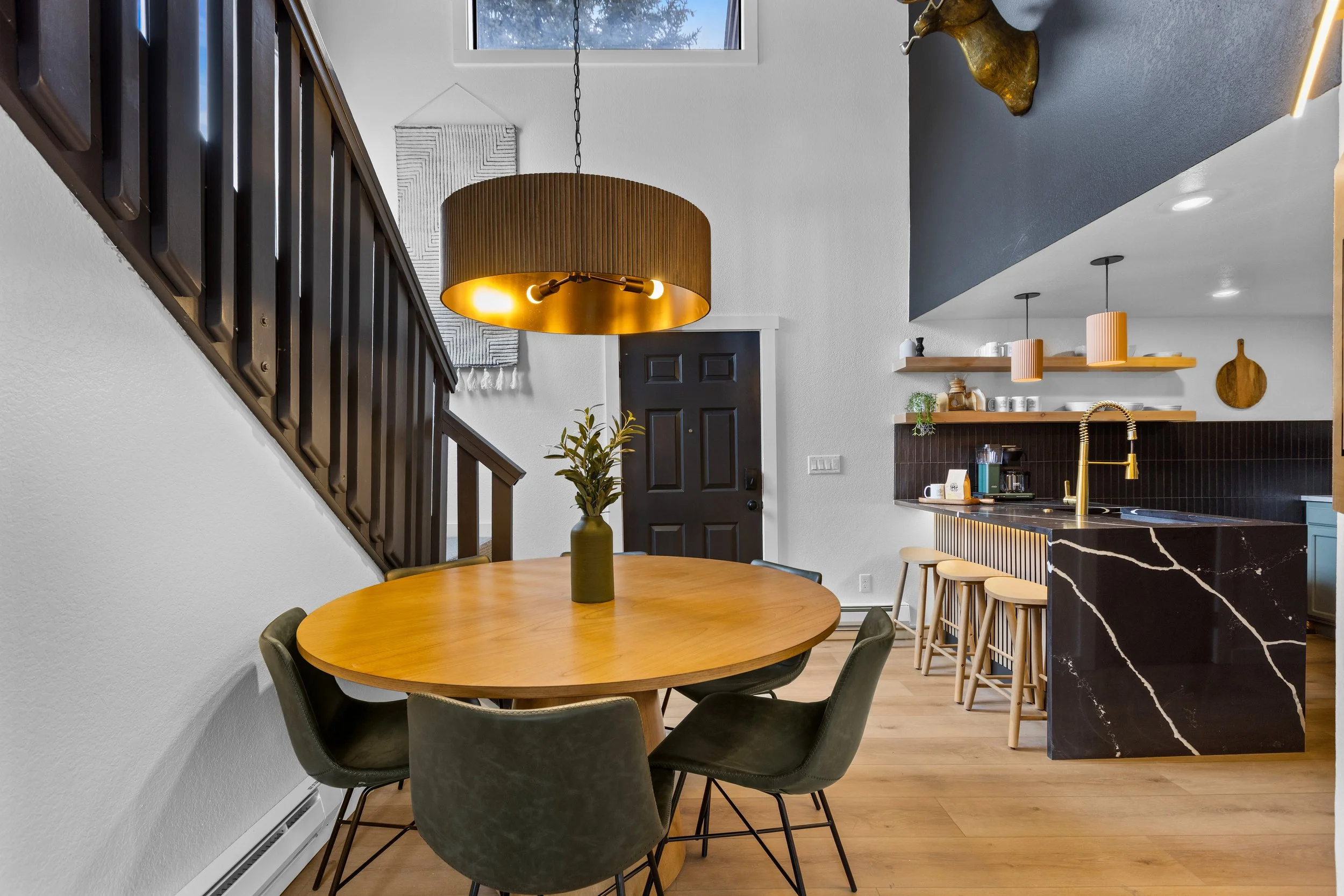 Interior view of a modern kitchen and dining area with a round wooden table, black chairs, a hanging pendant light, a dark front door, and open shelves with kitchen essentials.