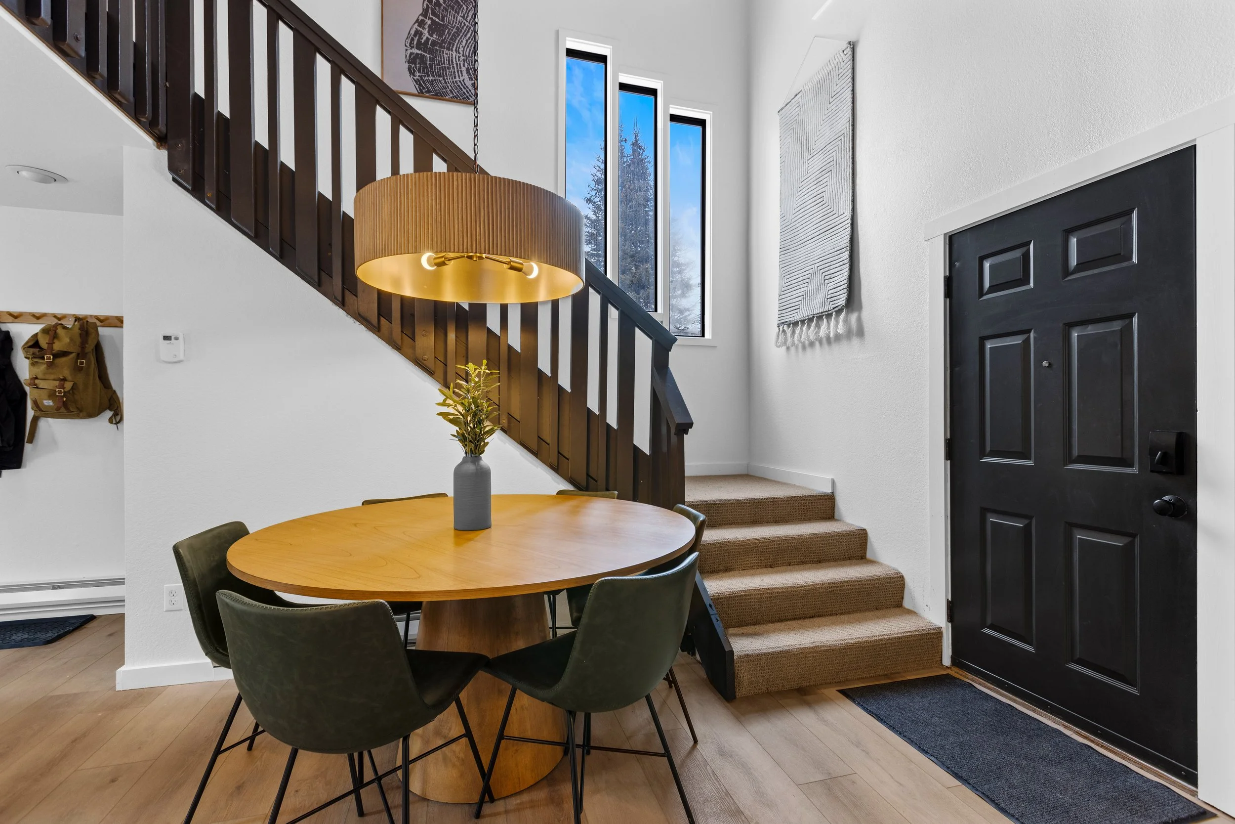Entryway with a black front door, a staircase with beige carpet, a wooden dining table with four green chairs, a hanging chandelier, a vase with a plant, windows showing a snowy landscape, and a wall hanging textile.