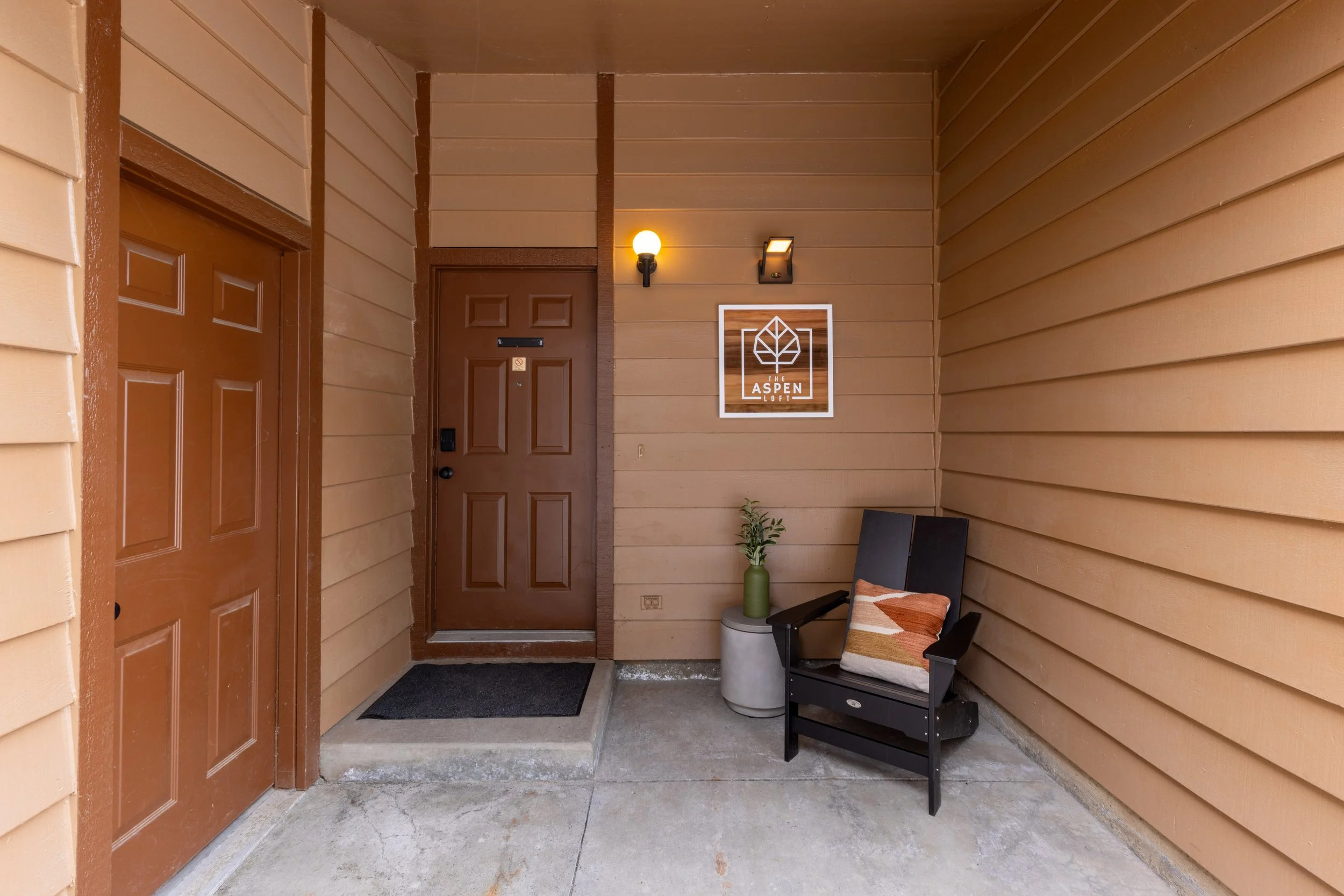 Apartment entrance with a brown door, a modern wall light, a decorative sign reading "The Aspen Loft," a small side table with a green vase and plant, and a black armchair with a decorative pillow, set against tan exterior walls.