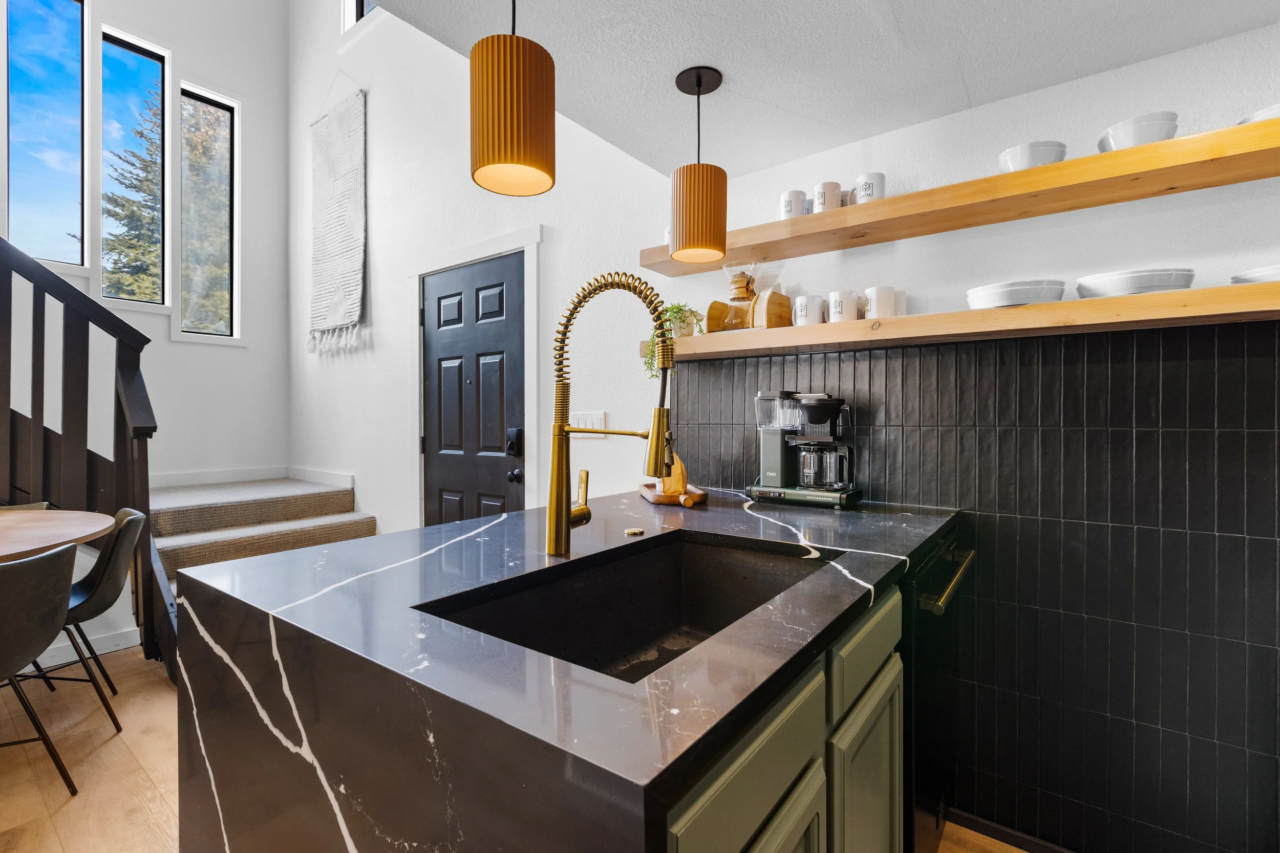 Modern kitchen with black marble island, gold faucet, black tiled wall, open wooden shelves with white dishware, and large windows showing blue sky and trees.