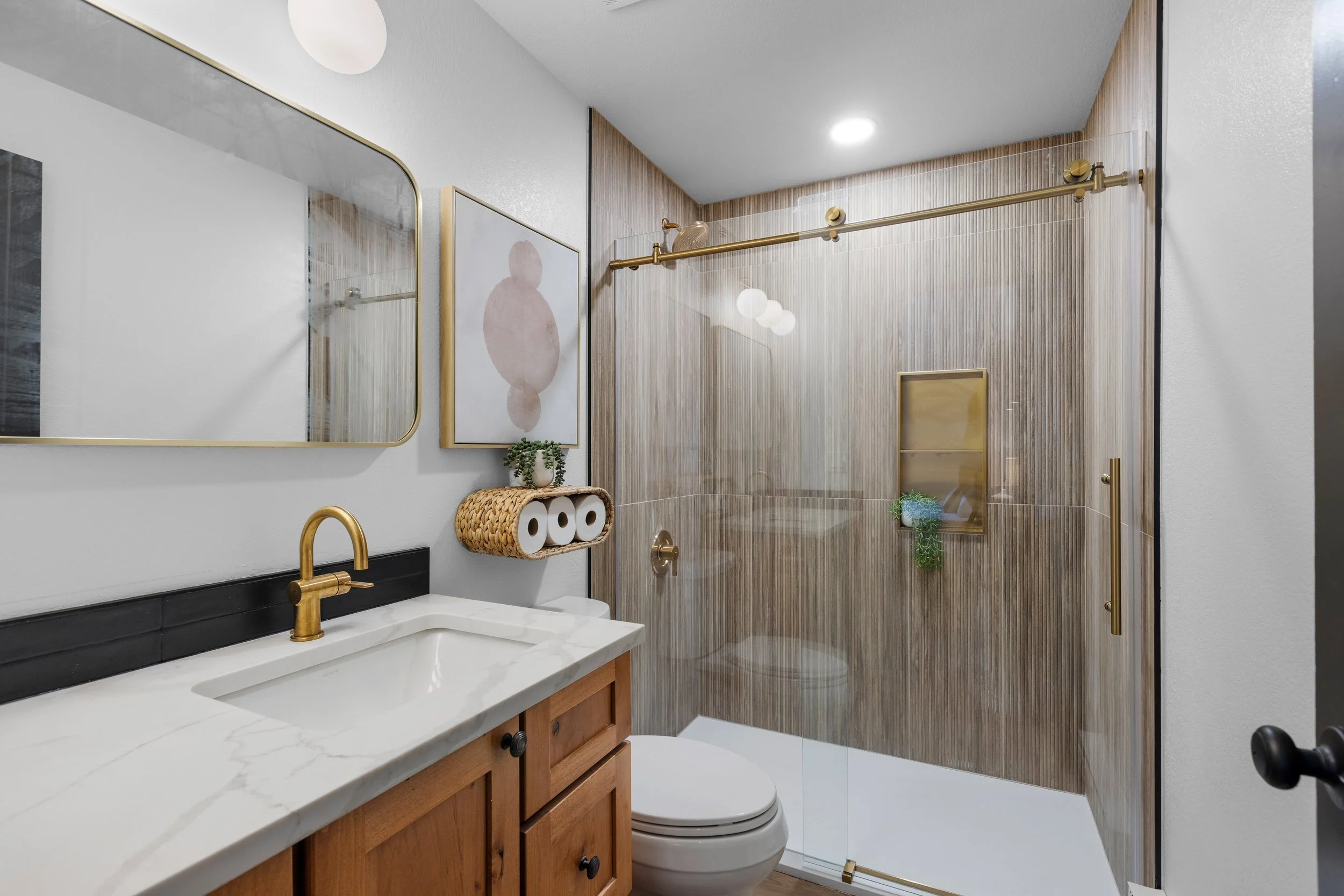 Bathroom with a wooden vanity, marble countertop, gold faucet, large mirror, framed artwork, toilet paper holder, and a glass shower with wood-look tile walls, gold fixtures, shower niche, and sliding glass door.