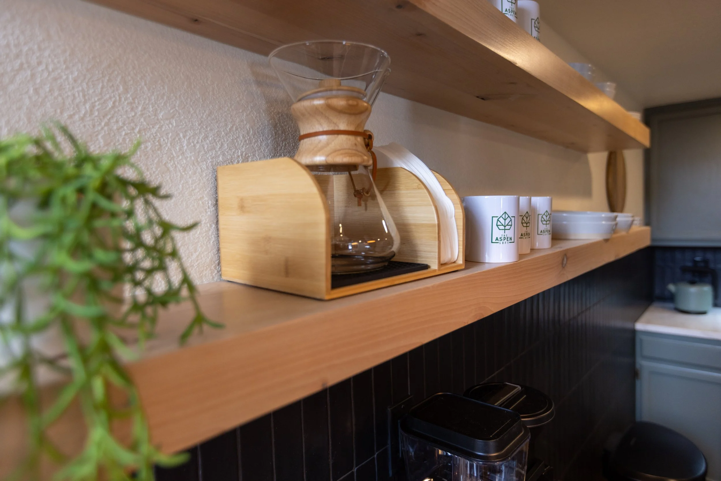 Wooden kitchen shelf holding a glass coffee carafe, white cups with a green leaf logo, and white bowls. A small green plant is partially visible on the left.