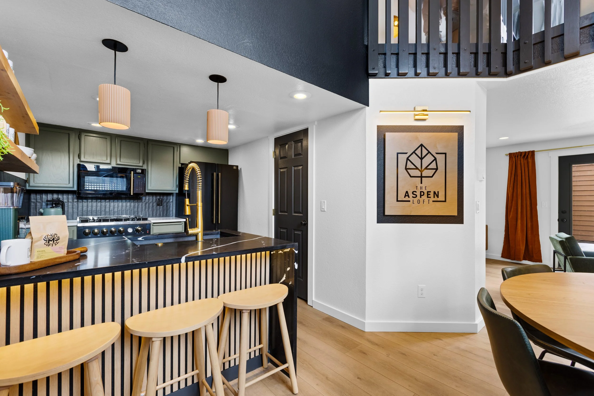Modern kitchen with gray cabinets, black appliances, a black and gold faucet, and a breakfast bar with three wooden stools. Adjacent dining area with a round table and black chairs, and a wall sign reading 'The Aspen Loft.'