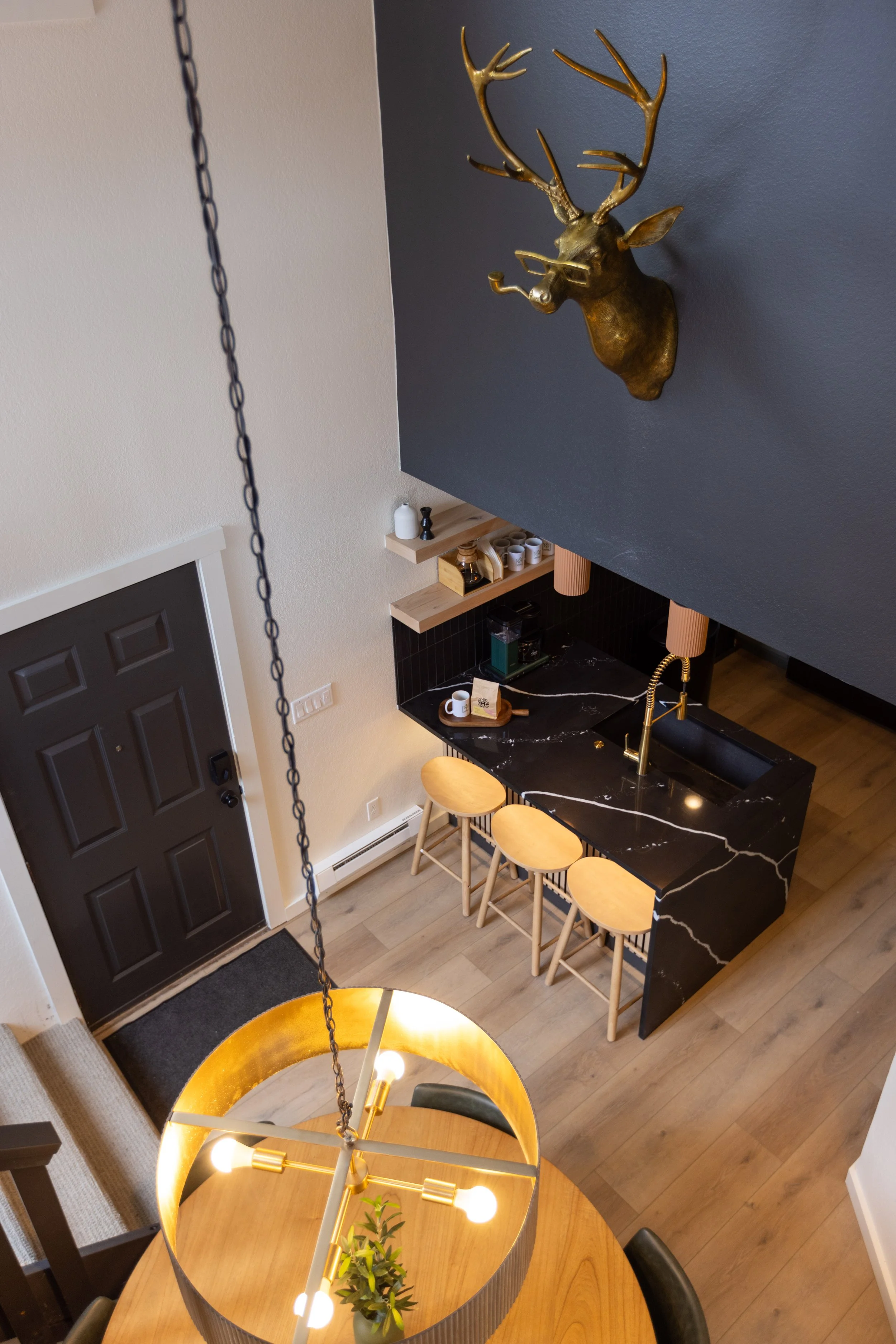 A top-down view of a modern kitchen with a black marble island, gold fixtures, a black deer head wall decoration, and a dining area with a gold chandelier and a wooden table with black chairs.