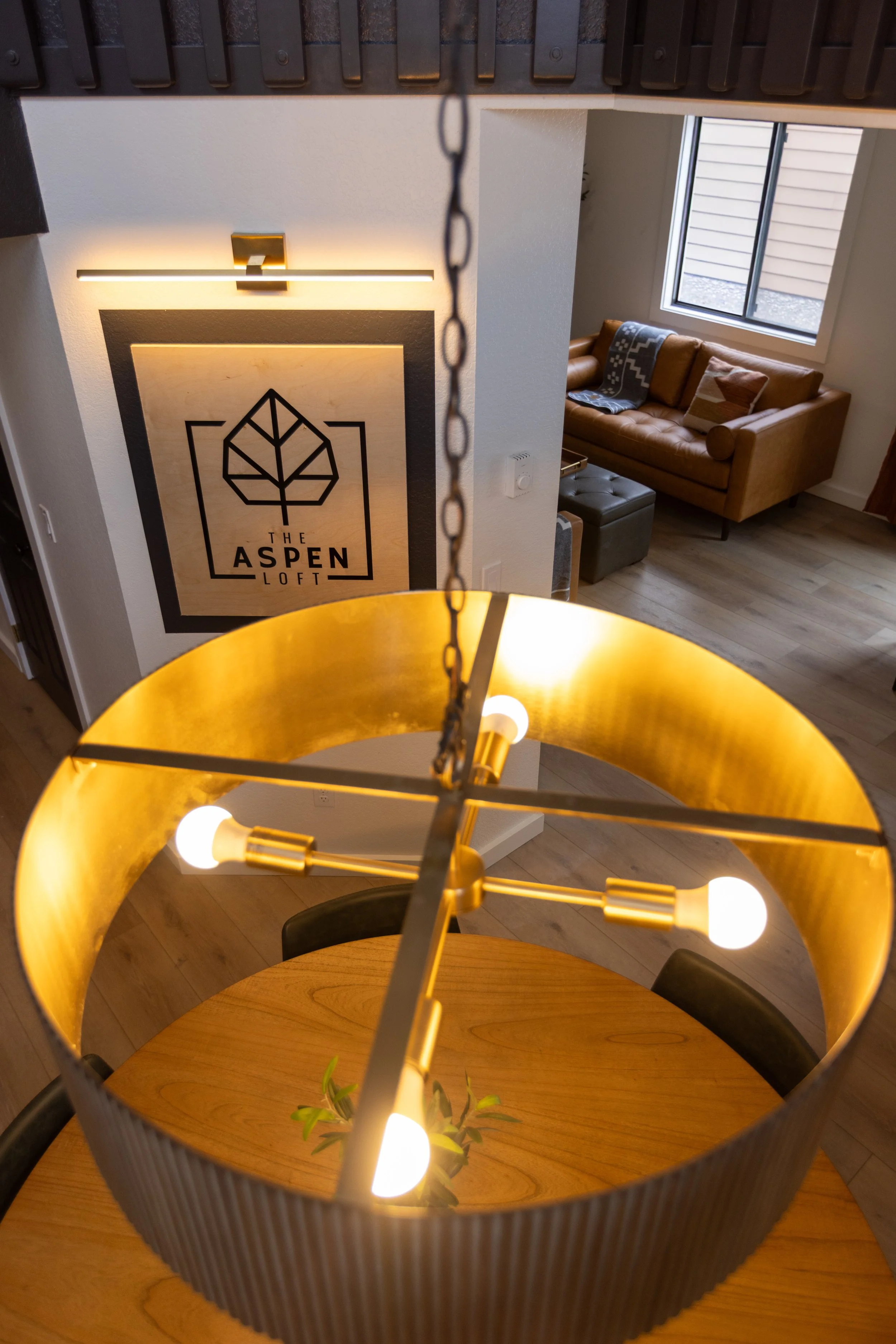 Interior view of a modern loft with a round dining table and a gold chandelier, a leather couch by a large window, and a wall sign reading 'The Aspen Loft' with a leaf logo.