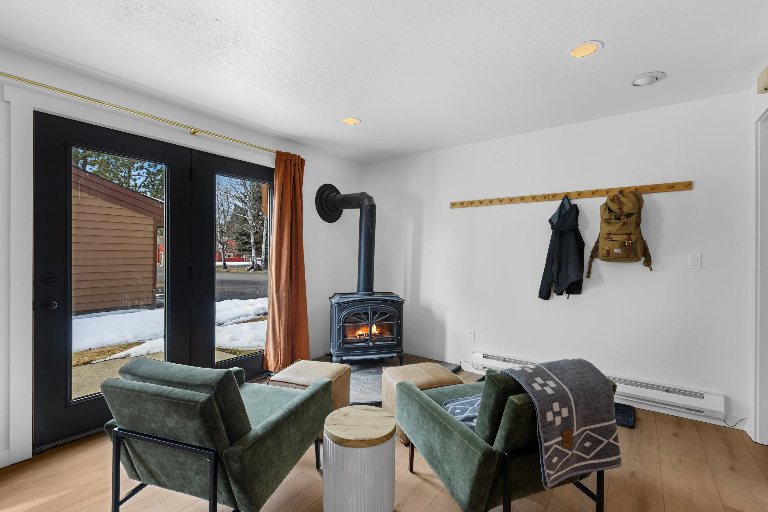 Living room with a wood stove, two green chairs, a small wooden table, and a sliding glass door with curtains showing snow outside.