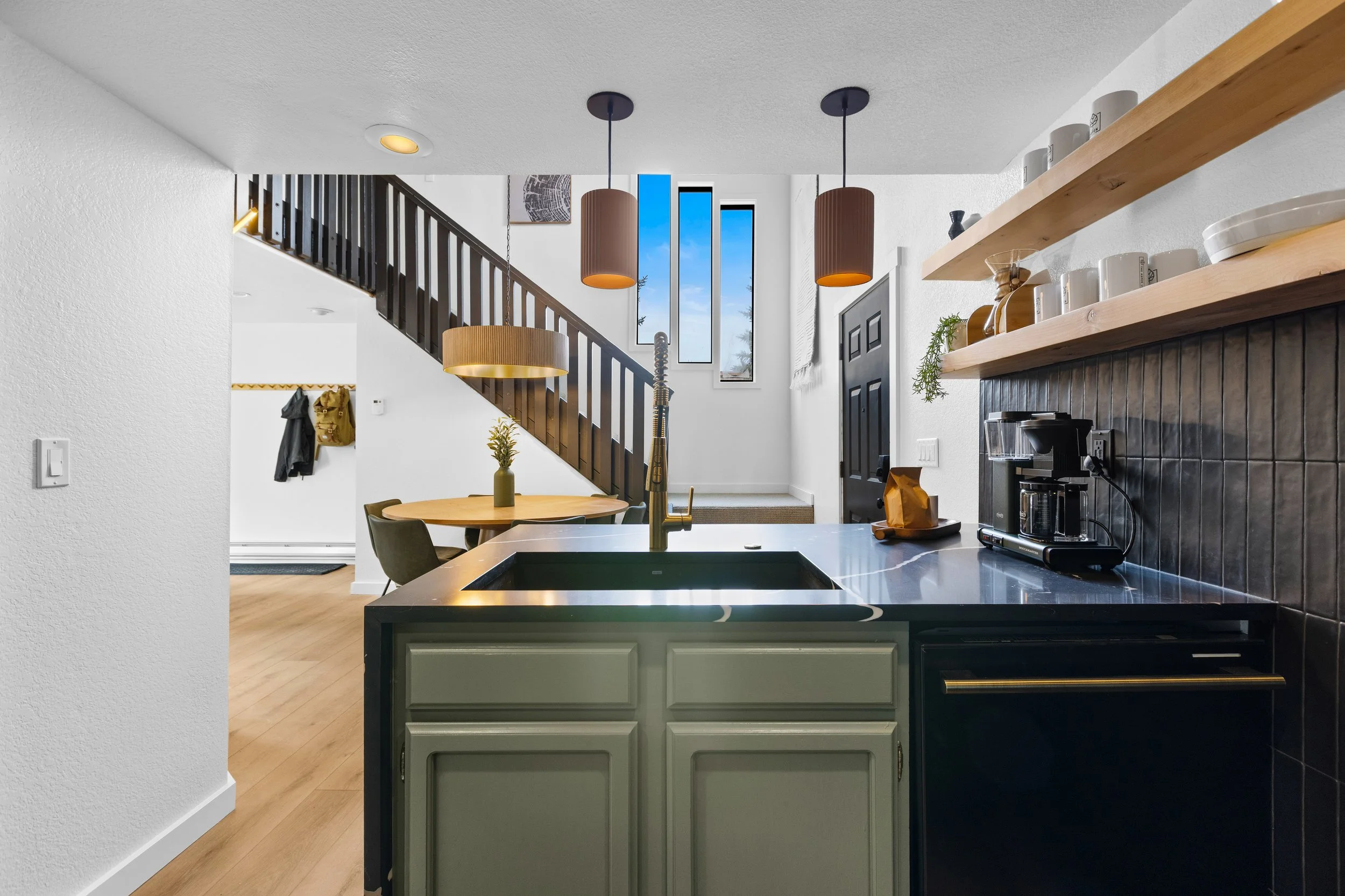 Modern kitchen with black countertop, white cabinets, and wooden open shelves holding dishes and decor. Black tile backsplash, coffee machine on counter, wooden dining table with green chairs, and three hanging pendant lights. Staircase with dark woo