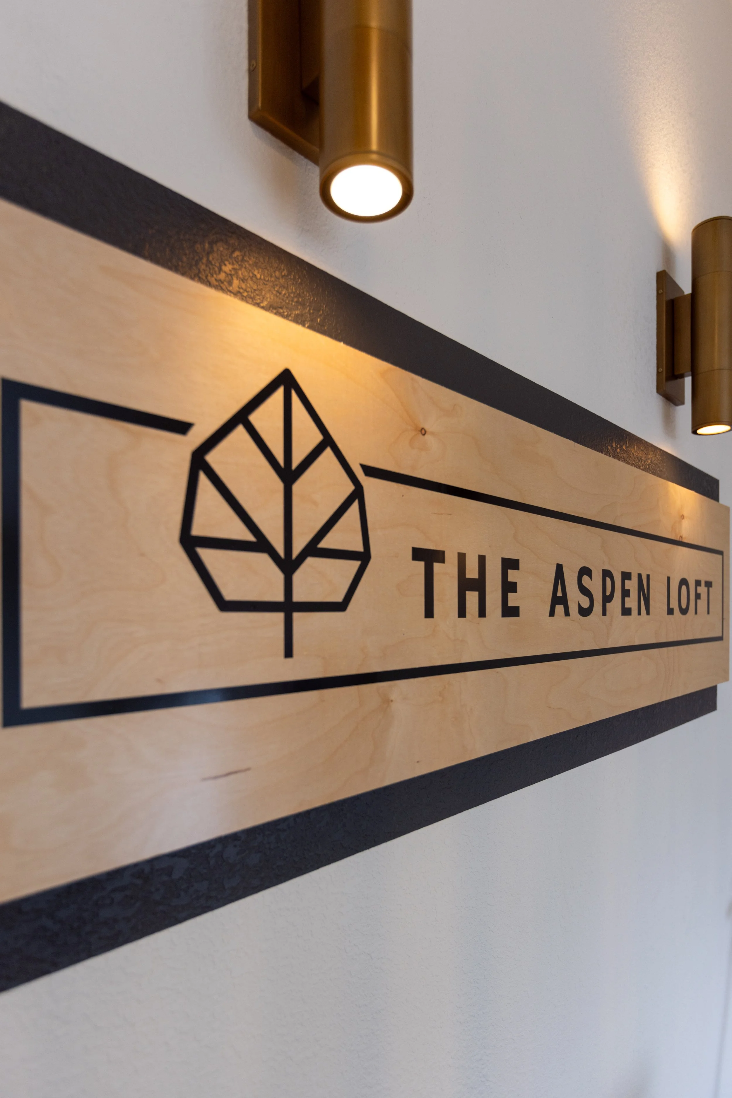 A wooden sign with a black geometric leaf graphic and the text 'The Aspen Loft,' mounted on a white wall with two gold-colored wall lights above.