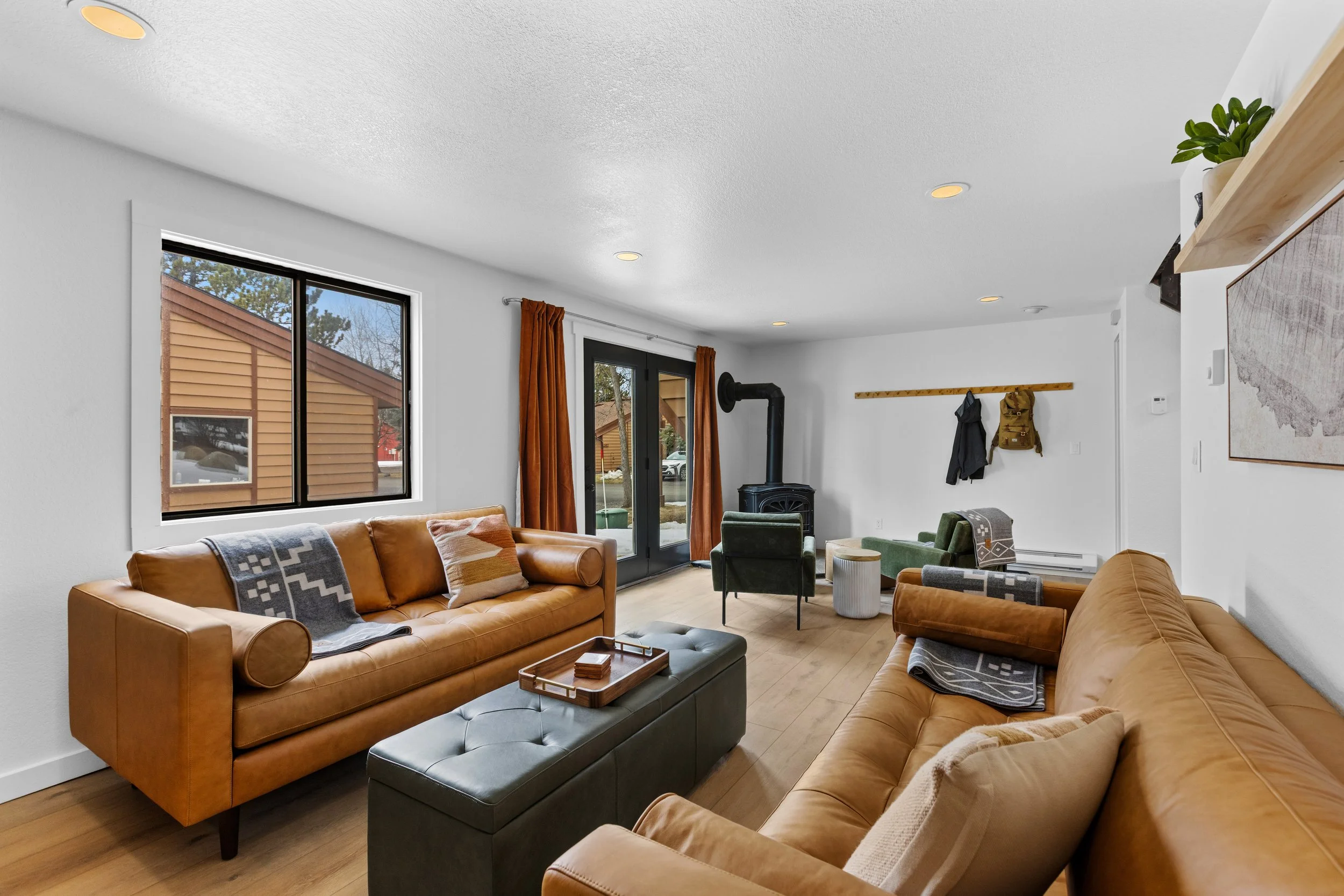 A cozy living room with two brown leather sofas, a green armchair, a black stove, a window, a sliding glass door, and a coat rack with jackets, featuring wooden flooring and white walls.