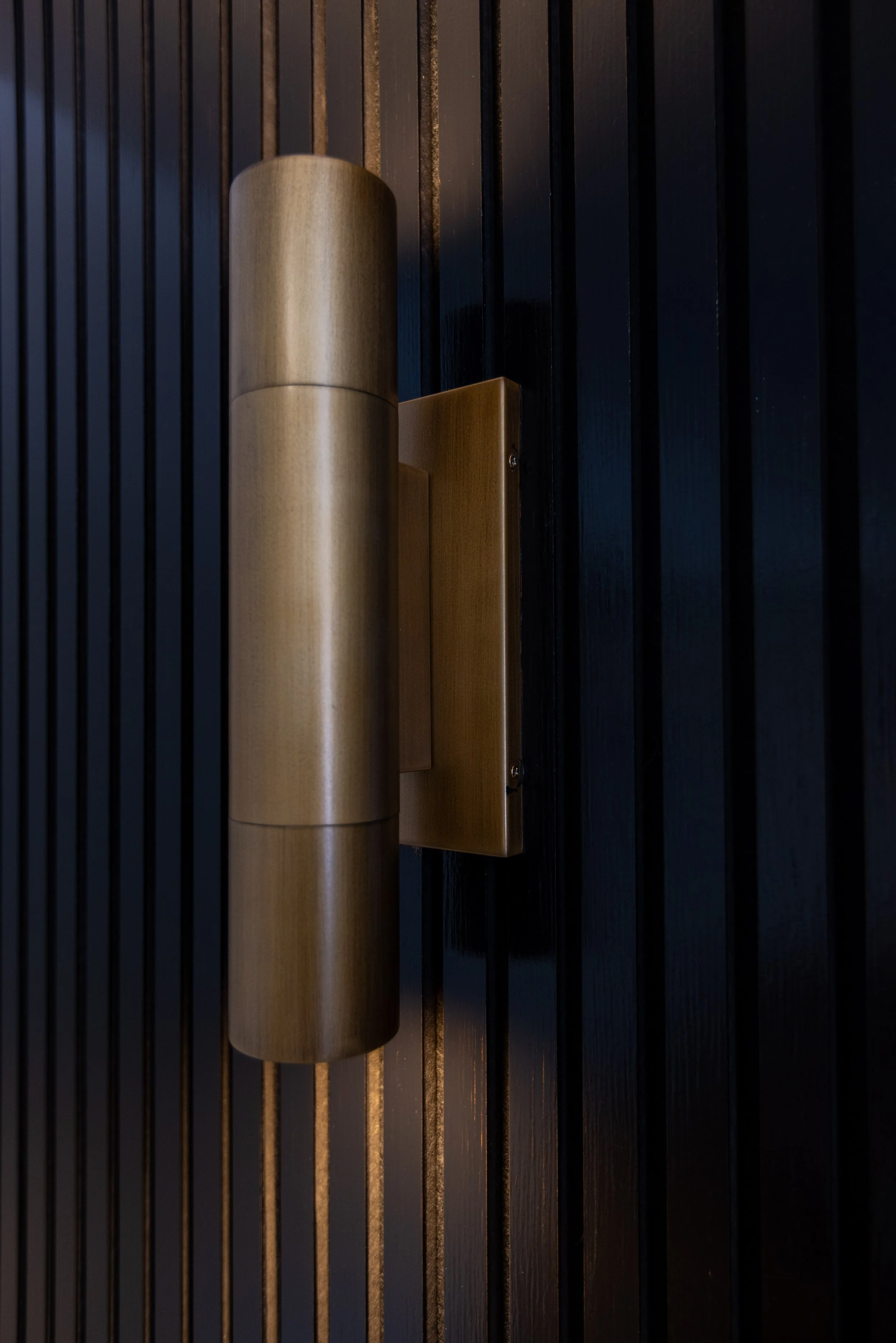 Close-up of a modern wall-mounted light fixture with a cylindrical gold-tone metal shade against a dark wooden slat wall.