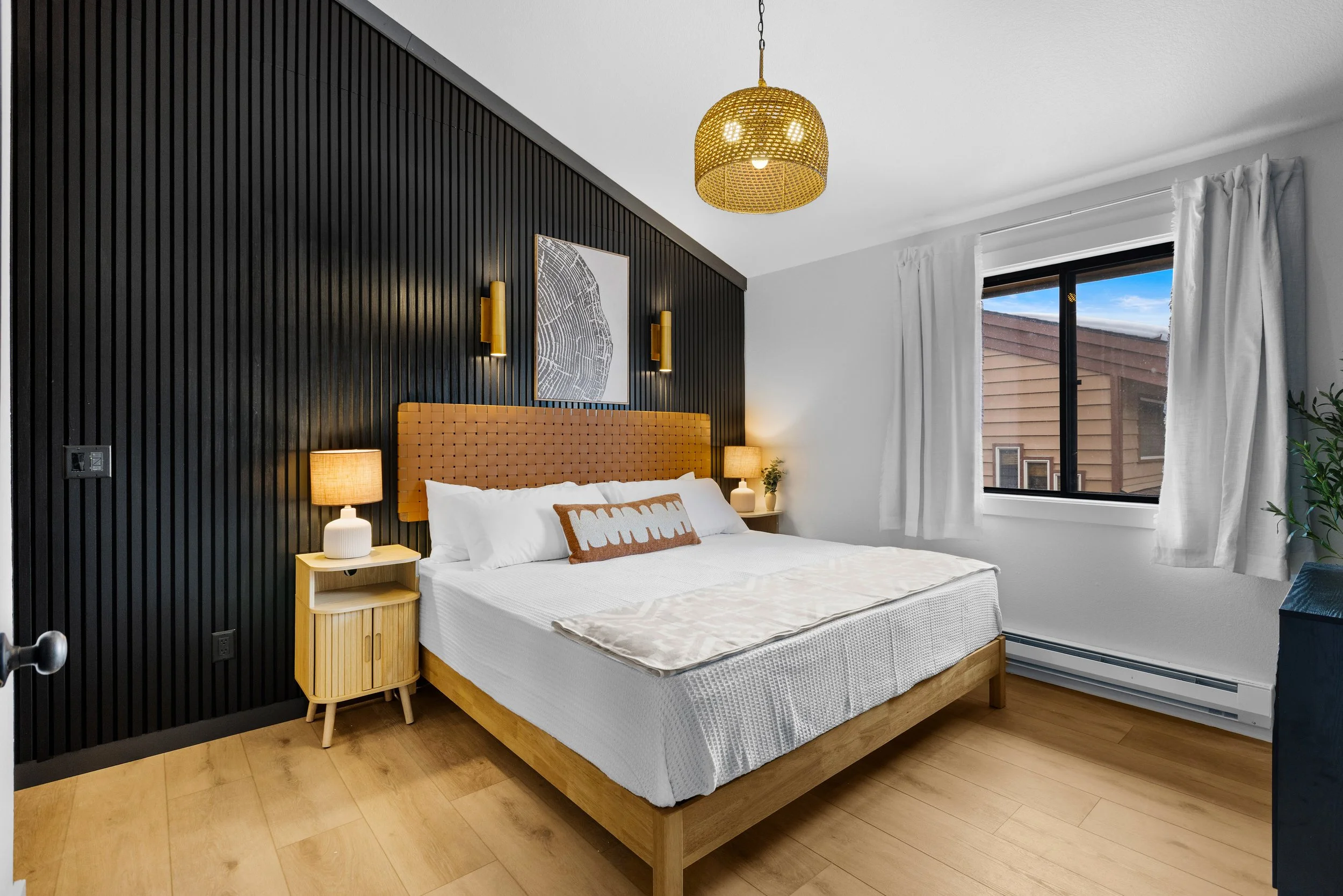 A modern bedroom with a black accent wall, tan headboard, white bedding, and wooden nightstands. The room has a window with white curtains and a gold ceiling light fixture.