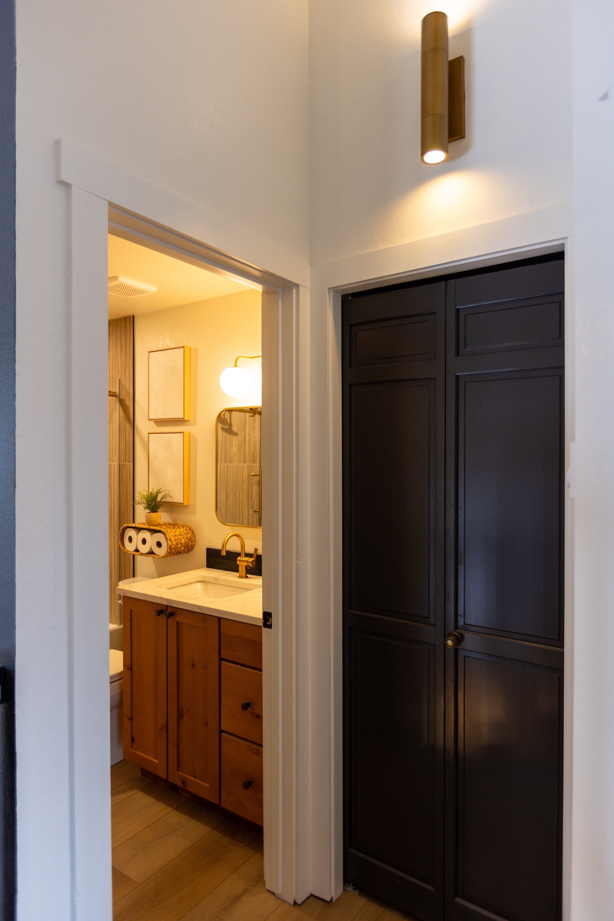 View of a bathroom through an open door with a wooden vanity, mirror, wall-mounted light fixture, and towels, with a dark cabinet in the foreground.