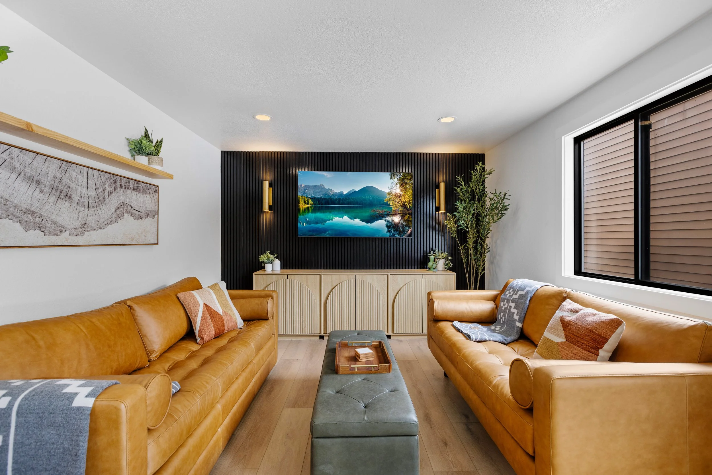 Living room with two tan leather sofas, a green ottoman, a wall-mounted TV showing a mountain lake scene, a black accent wall, large window with brown blinds, and various plants and decorative items.