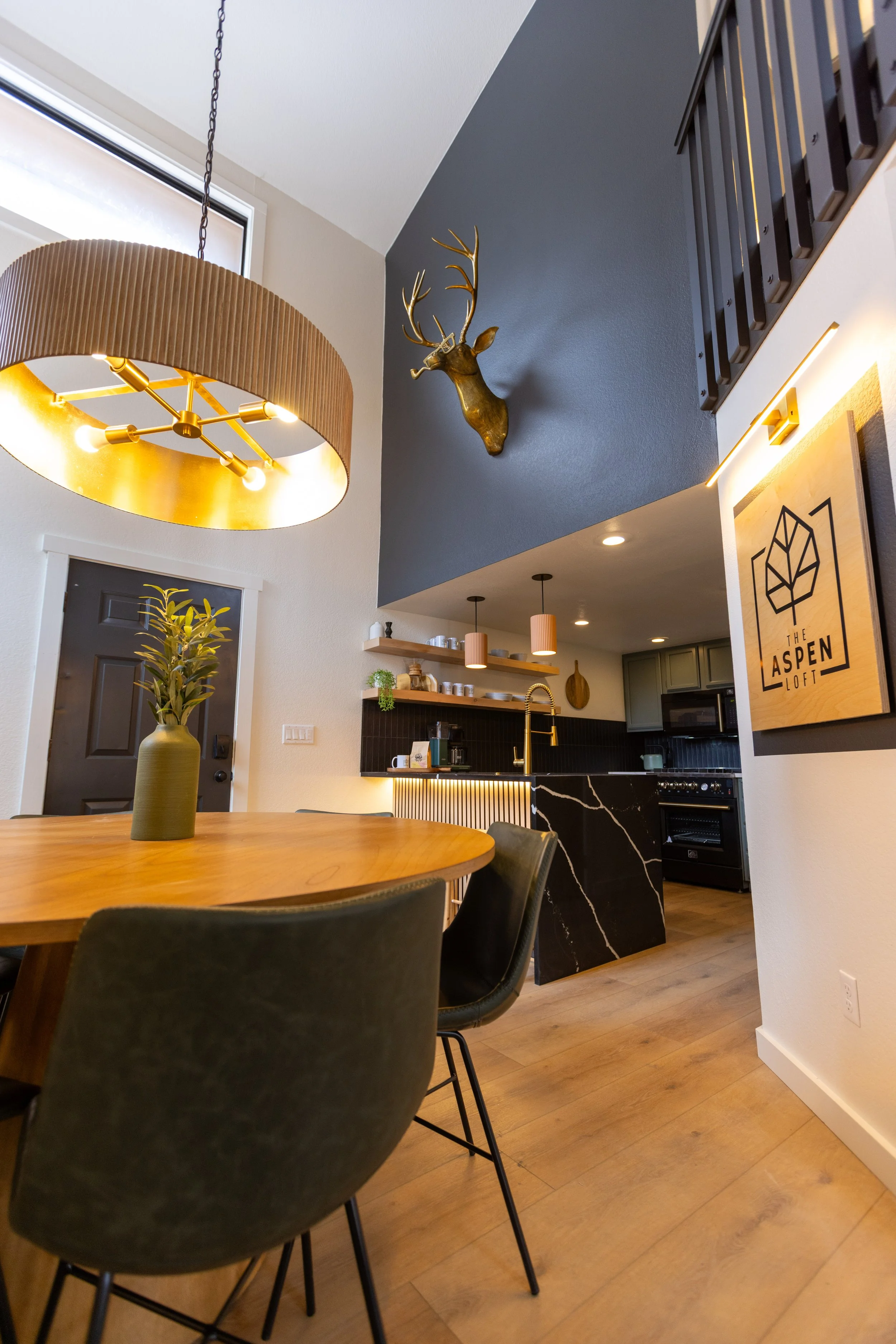 Interior view of a modern kitchen and dining area with a round wooden table, black chairs, a black marble kitchen island, and a ceiling light fixture. A mounted deer head is on the blue accent wall, and a framed art piece is on the white wall. The sp