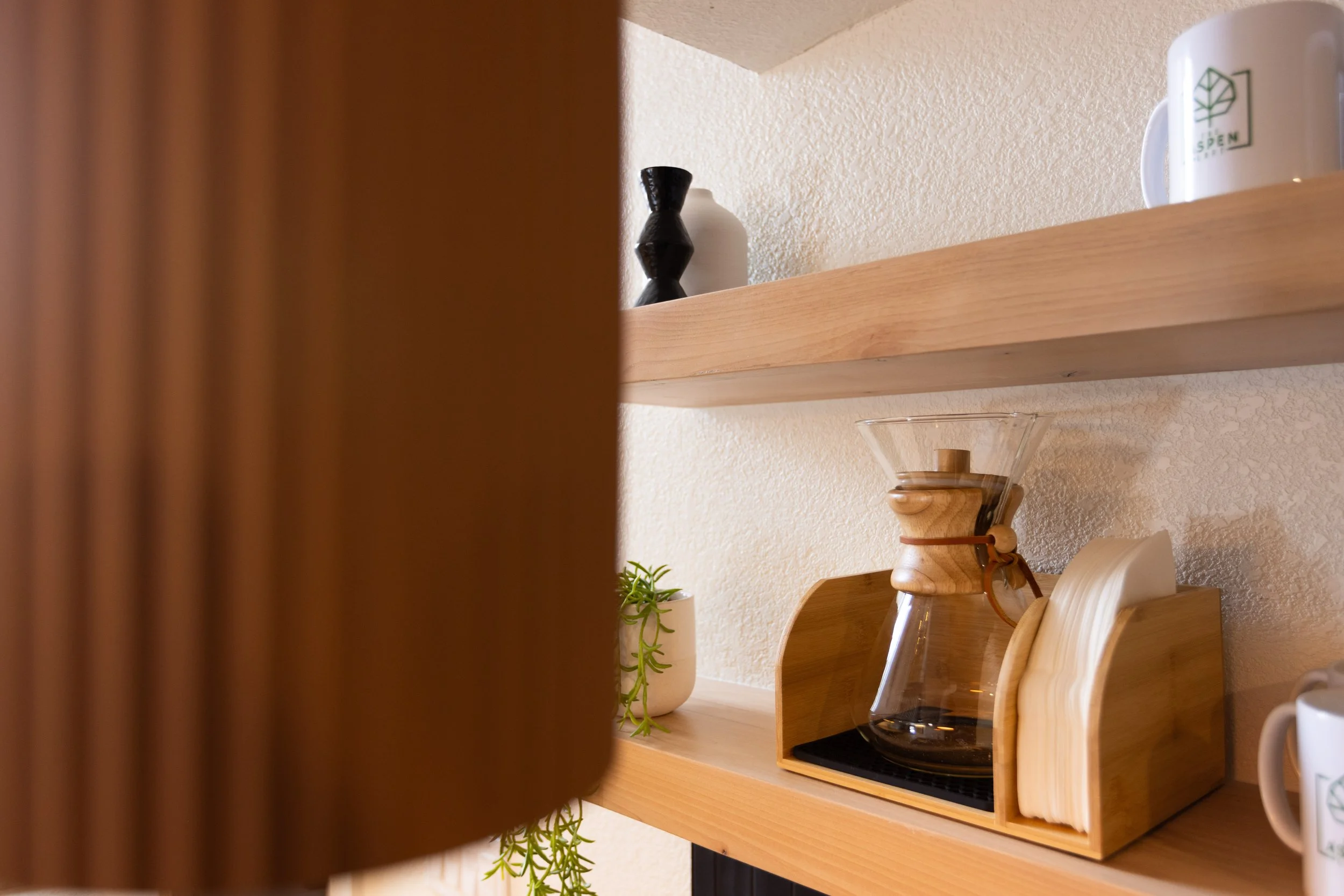 Coffee-making setup on wooden shelves with a glass pour-over coffee maker, paper filter holder, small plant, and decorative vases.