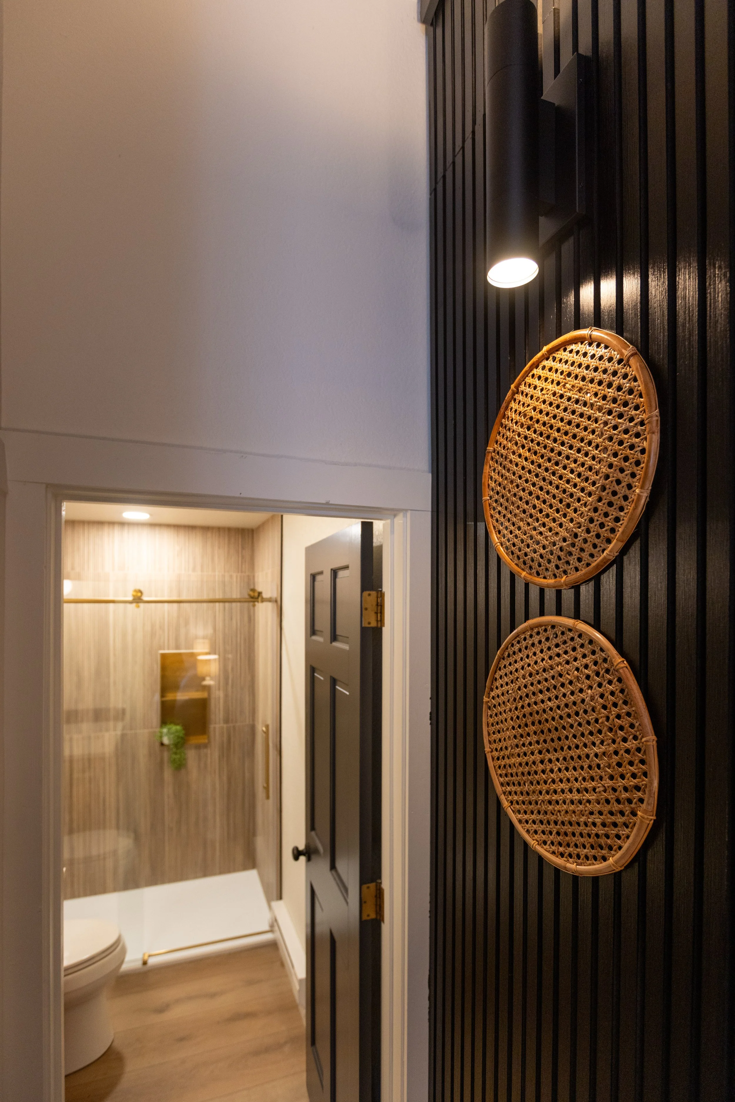 A hallway with black vertical paneling on the wall, a black ceiling-mounted light fixture, and two round wicker serving trays hanging on the wall. An open door shows a bathroom with wood-look tile on the walls, a toilet, and a mirror with potted plan