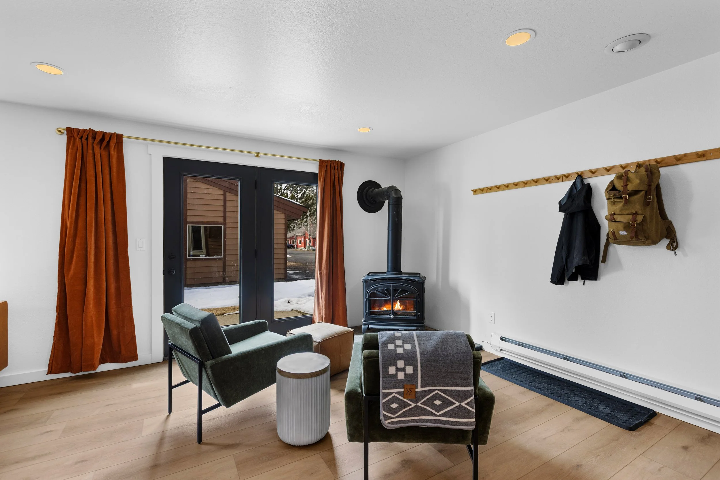 Living room with a wood stove, black door with glass panes, rust-colored curtains, two green armchairs, a small white side table, a gray patterned blanket, and a wall-mounted coat rack with jackets and a backpack.