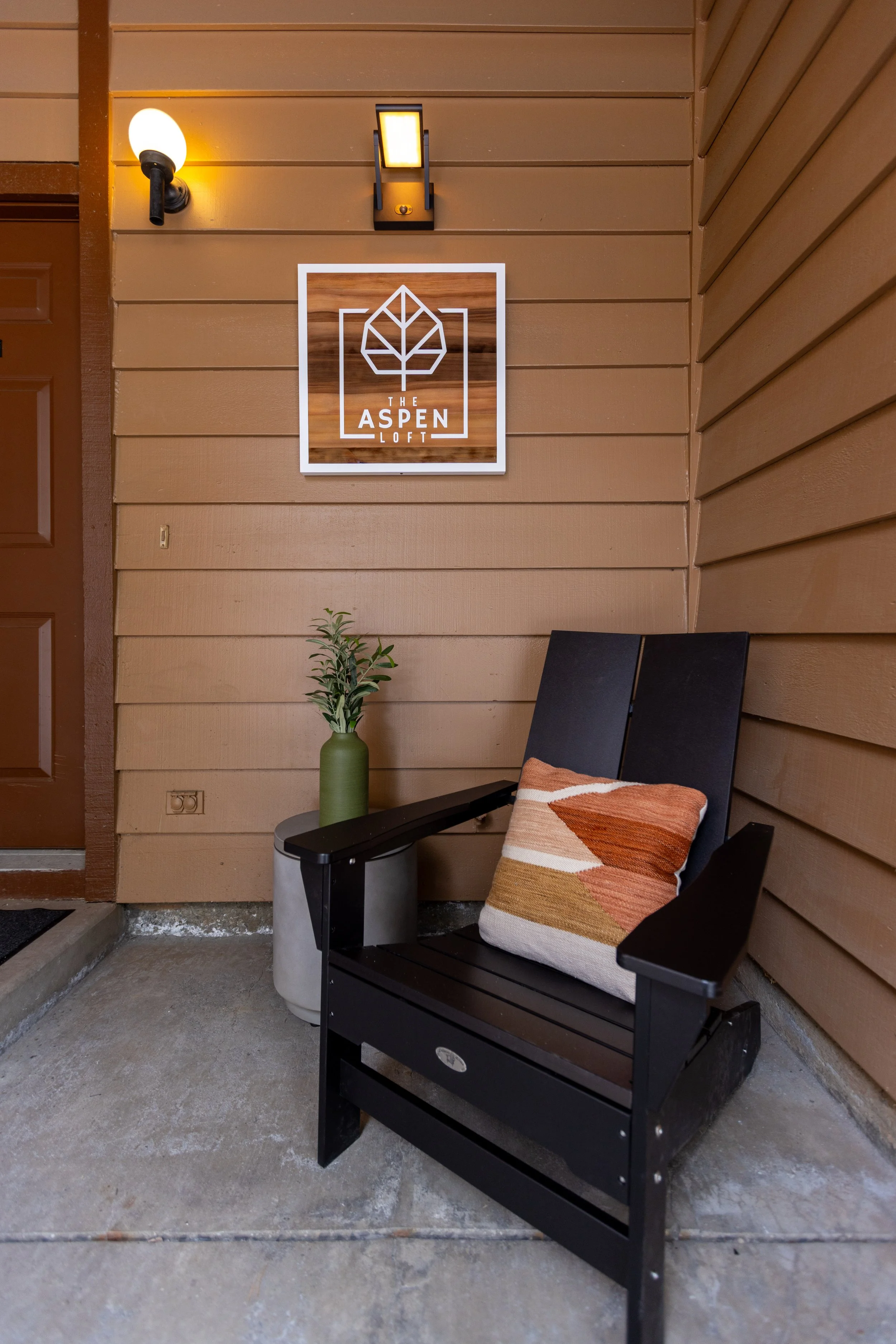 A black outdoor rocking chair with a patterned cushion, positioned next to a green vase with plant, on a concrete porch, in front of a wooden wall with a sign that reads 'The Aspen Loft'.