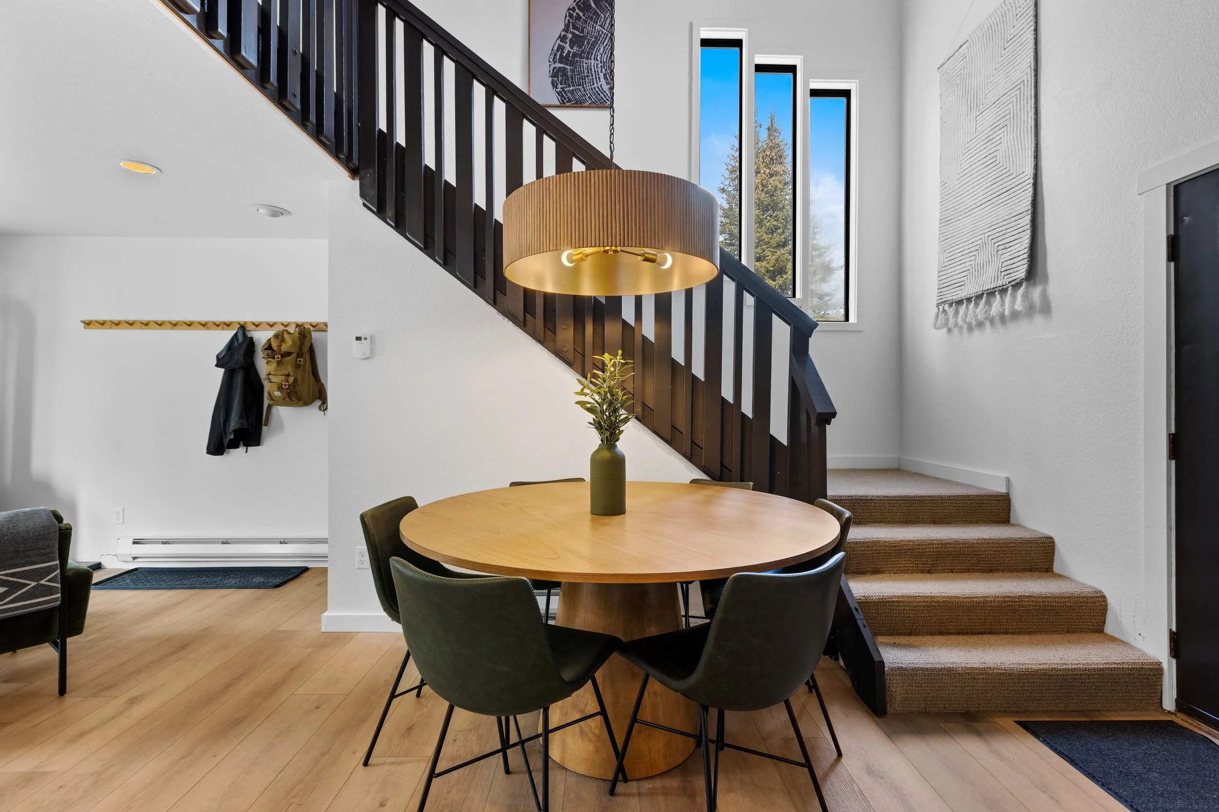 Interior of a modern home with a round wooden dining table, five black chairs, a large pendant light, a staircase with dark railing, and a wall with tall windows showing trees outside.