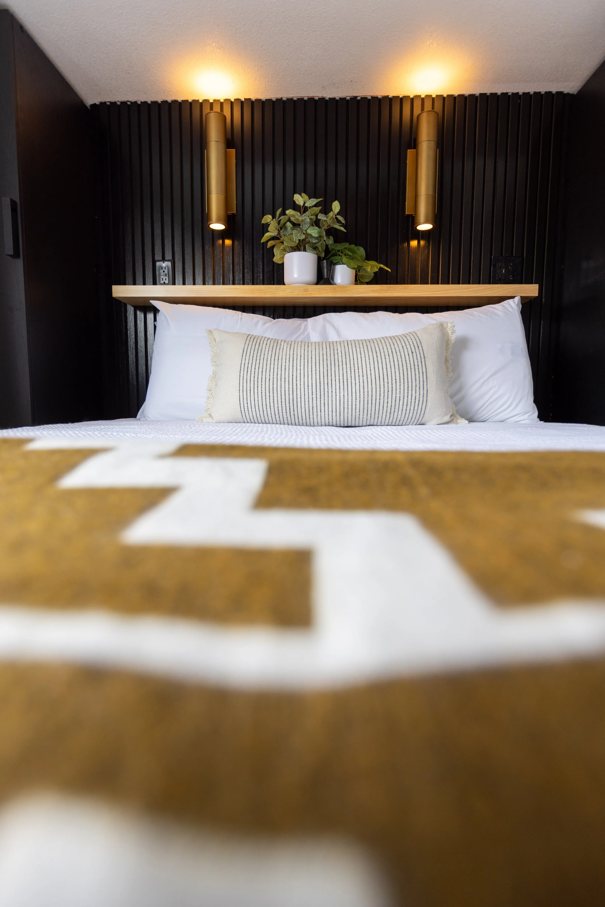 A bedroom with a bed, white pillows, a beige decorative pillow, and a geometric-patterned blanket. Above the bed is a wooden shelf with potted plants. Two vertical gold wall sconces are mounted on a black slatted wood headboard, with warm lights illu
