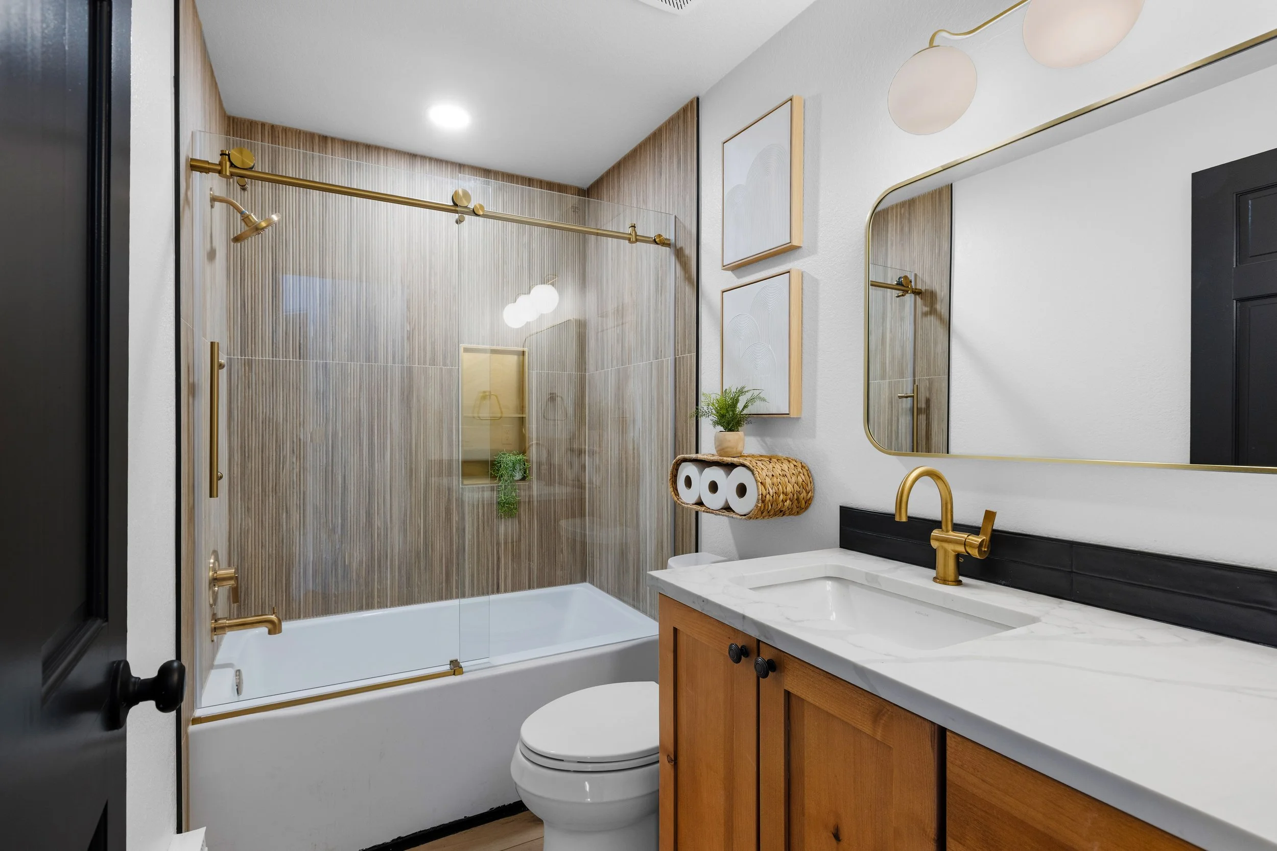 Modern bathroom with a bathtub and shower, wooden vanity with white marble countertop, gold fixtures, mirror, framed art, and toilet.
