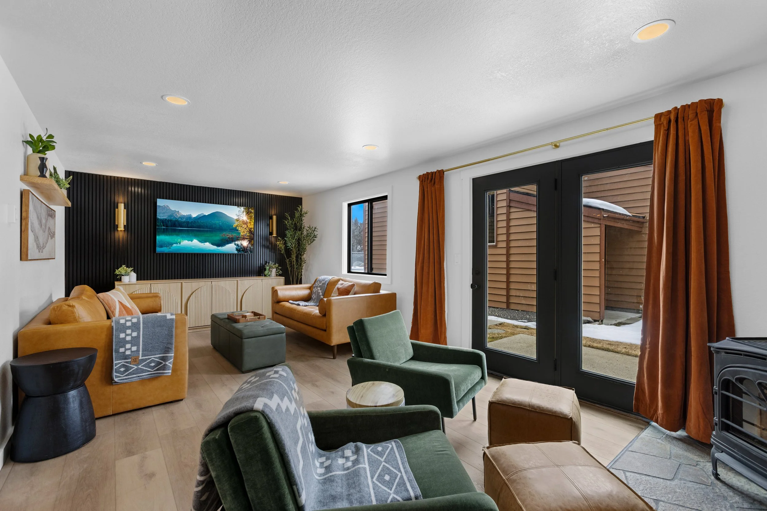 Living room with two mustard-colored sofas, green armchairs, a wall-mounted TV, and large windows with orange curtains.